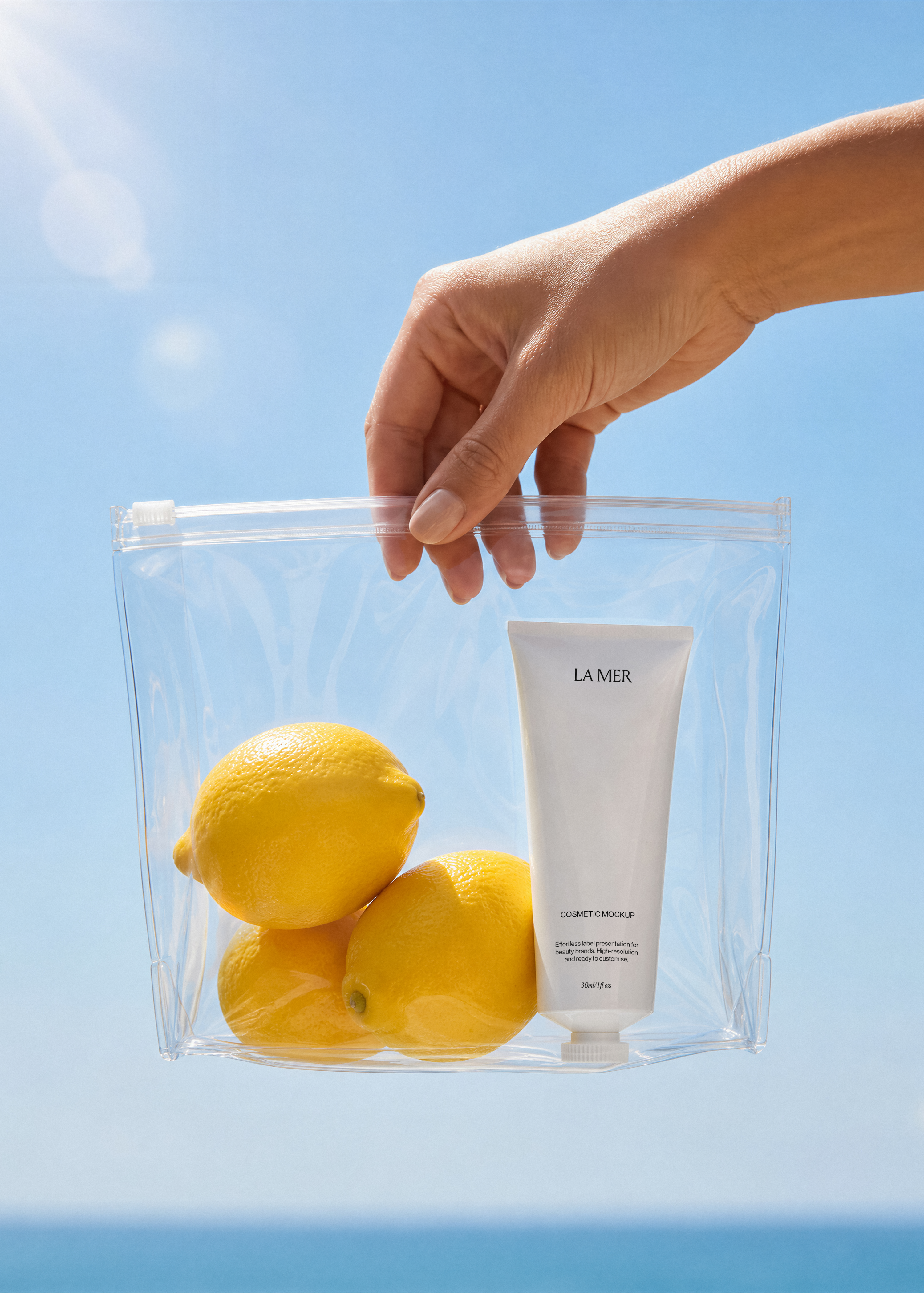 White cosmetic tube mockup and three lemons inside a clear zip pouch held by a hand against a blue sky, coastal styling