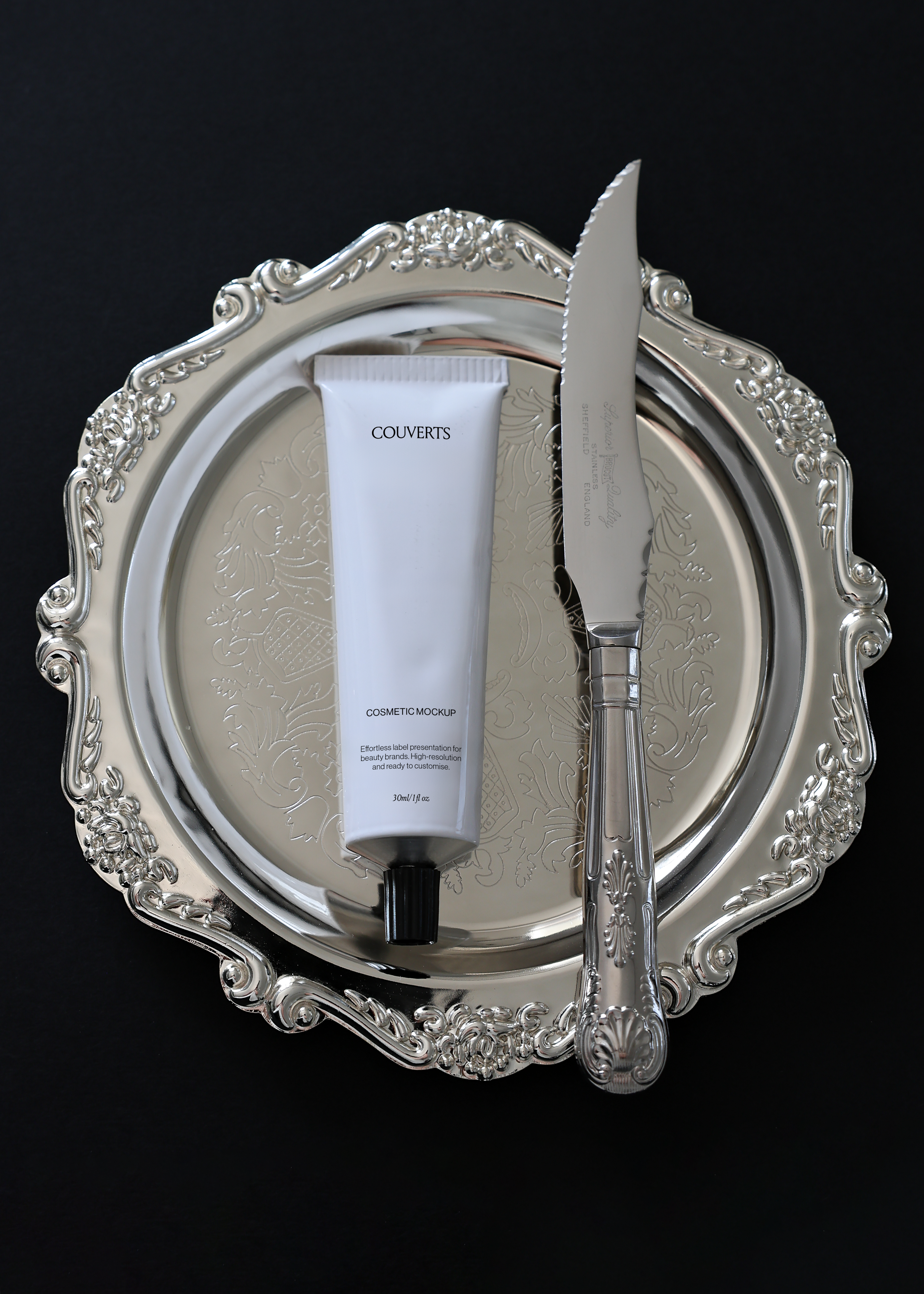 Cosmetic tube mockup on ornate silver dinner plate with serrated knife, black background, vintage tableware styling