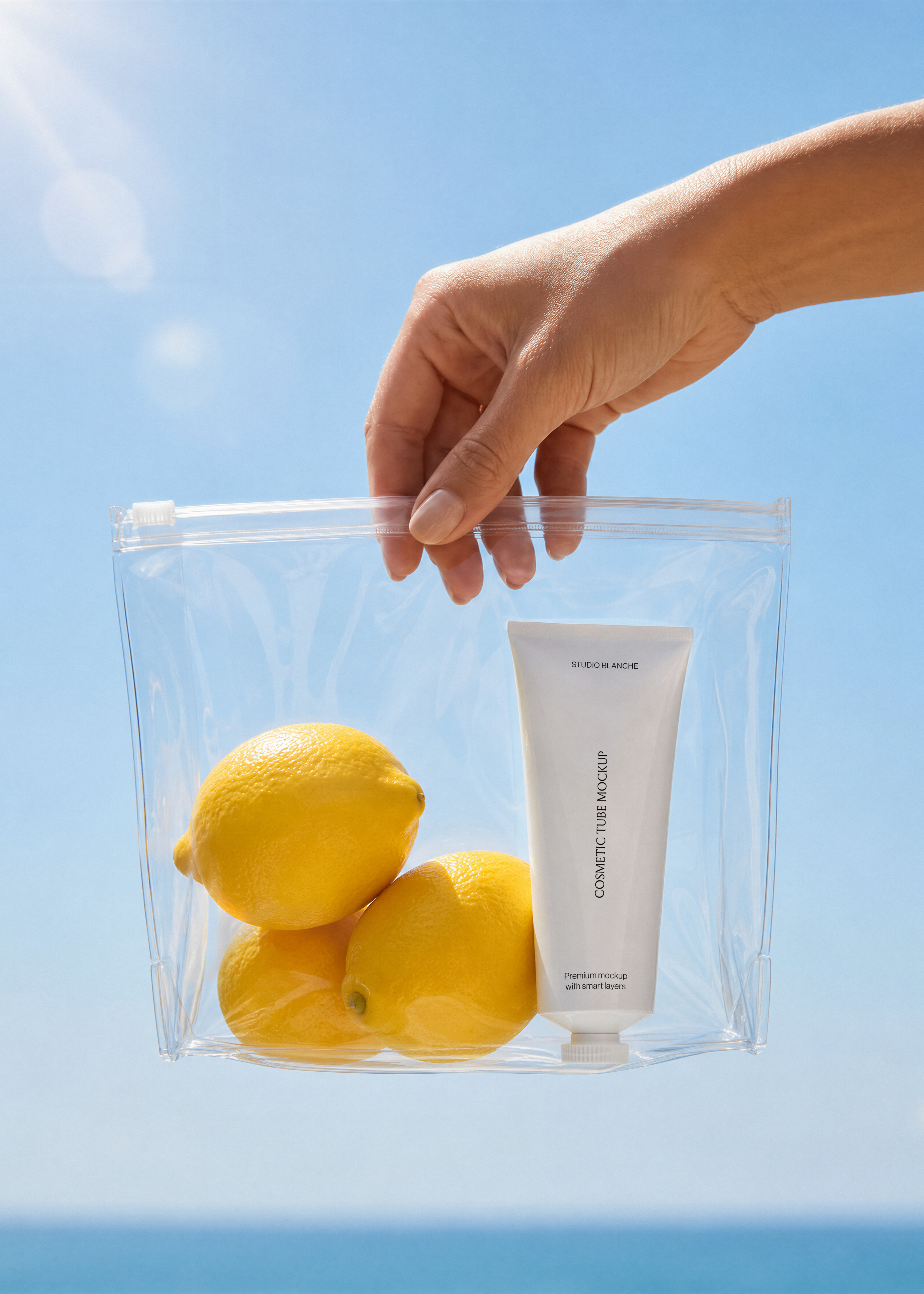 Close view of a white cosmetic tube label in a transparent pouch beside lemons, blue sky background, minimal coastal scene