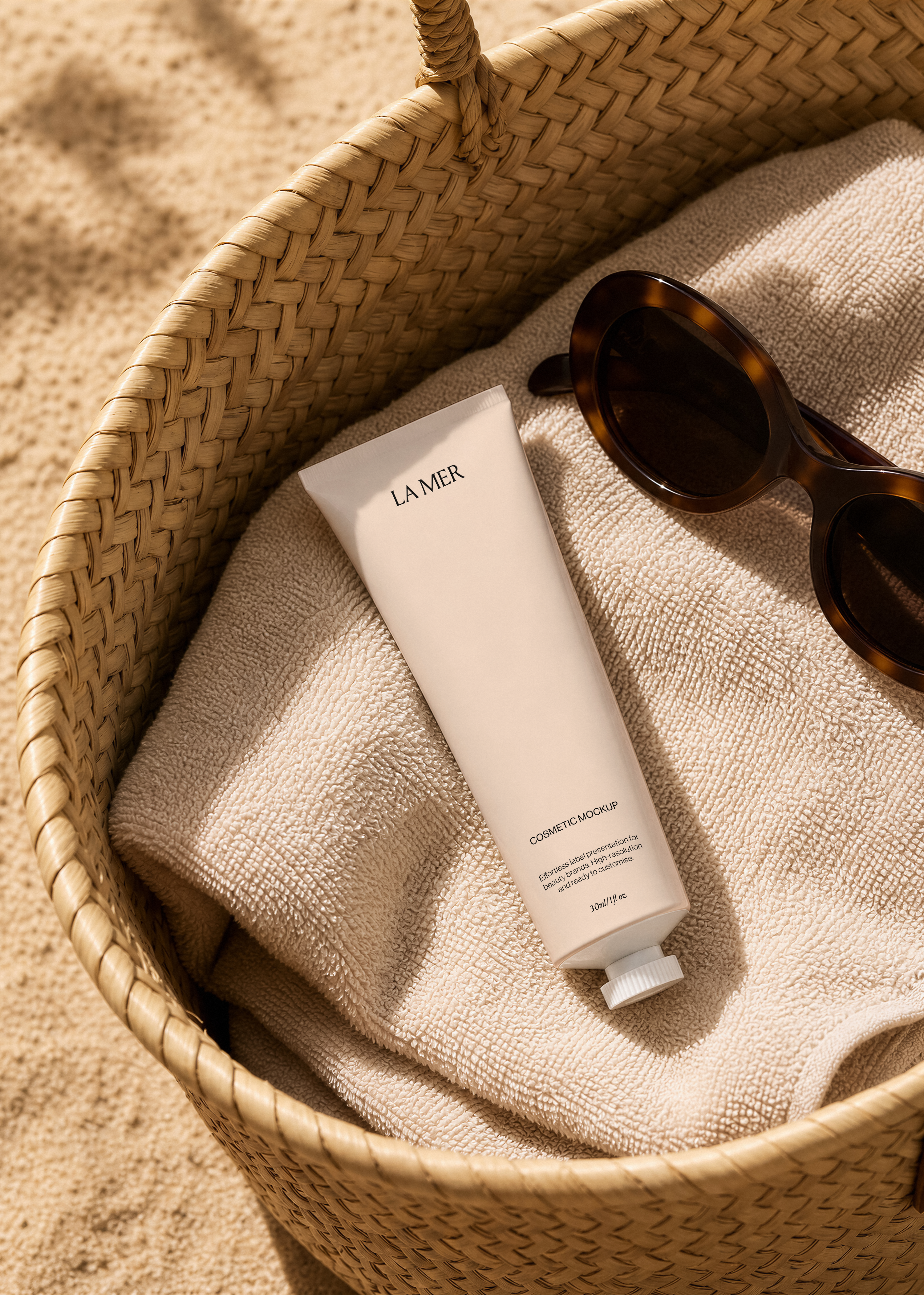 White cosmetic tube mockup on a beige towel inside a woven straw beach bag with tortoiseshell sunglasses on sand