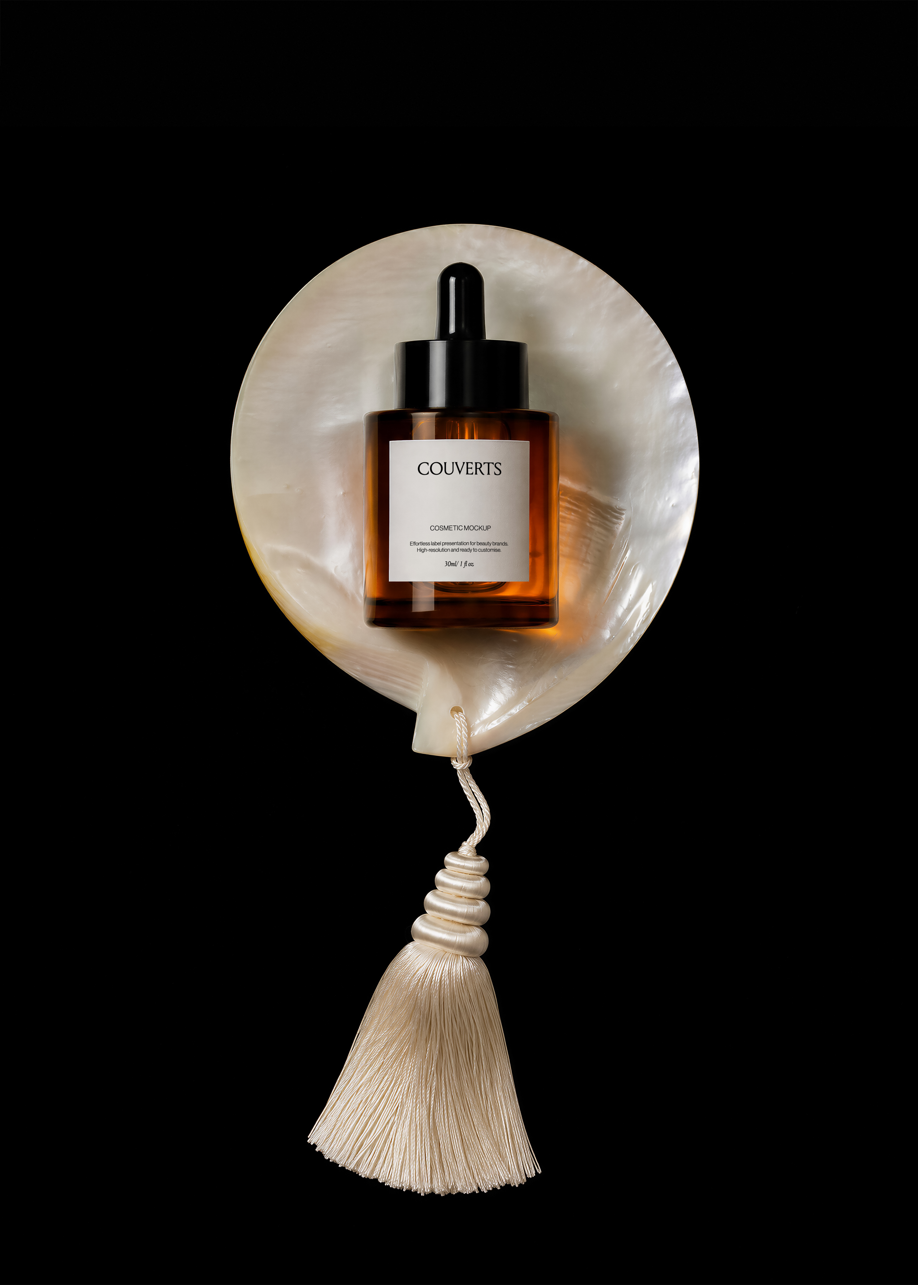 Amber glass dropper bottle mockup with black cap resting on a mother-of-pearl shell with cream silk tassel on black
