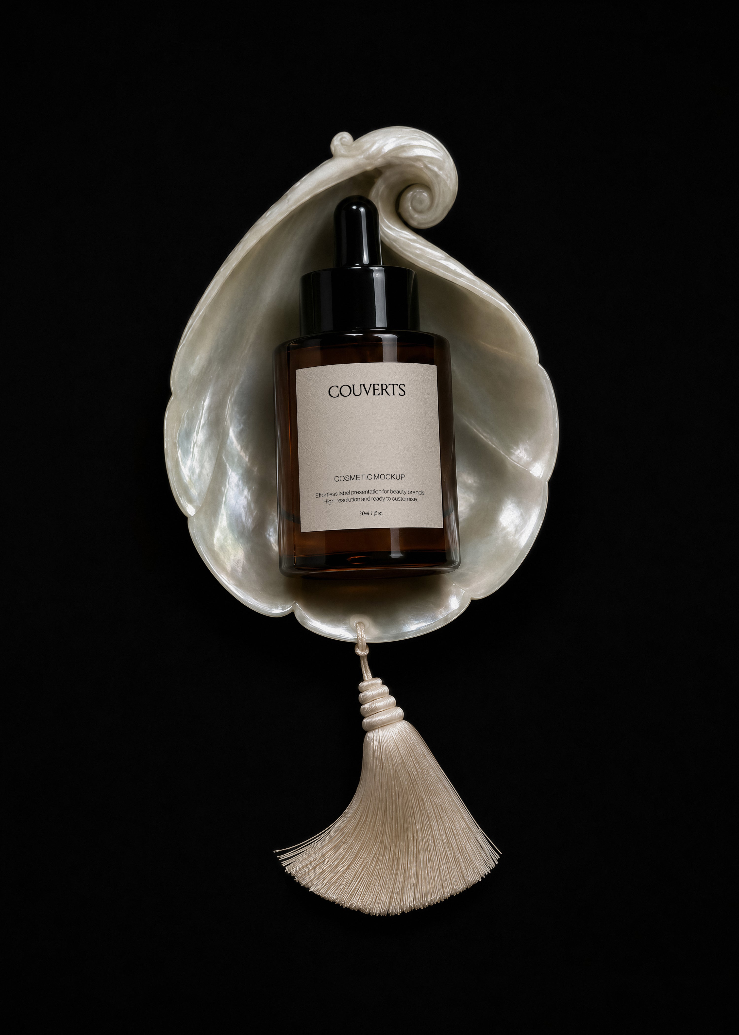 Amber glass dropper bottle mockup with black cap resting on a mother-of-pearl shell with cream silk tassel on black