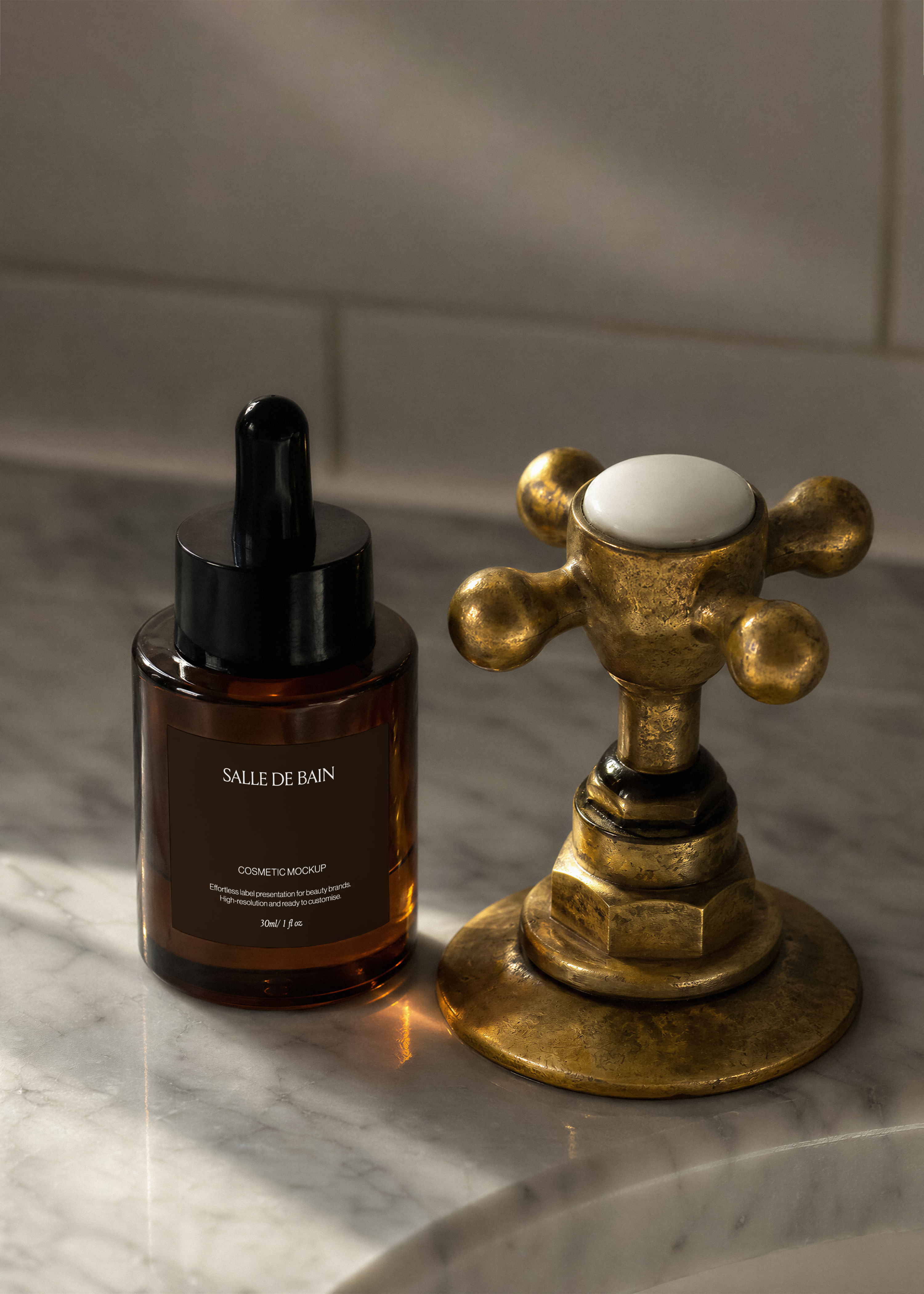 Amber dropper bottle mockup with black cap on white marble beside aged brass cross-handle tap, warm directional light