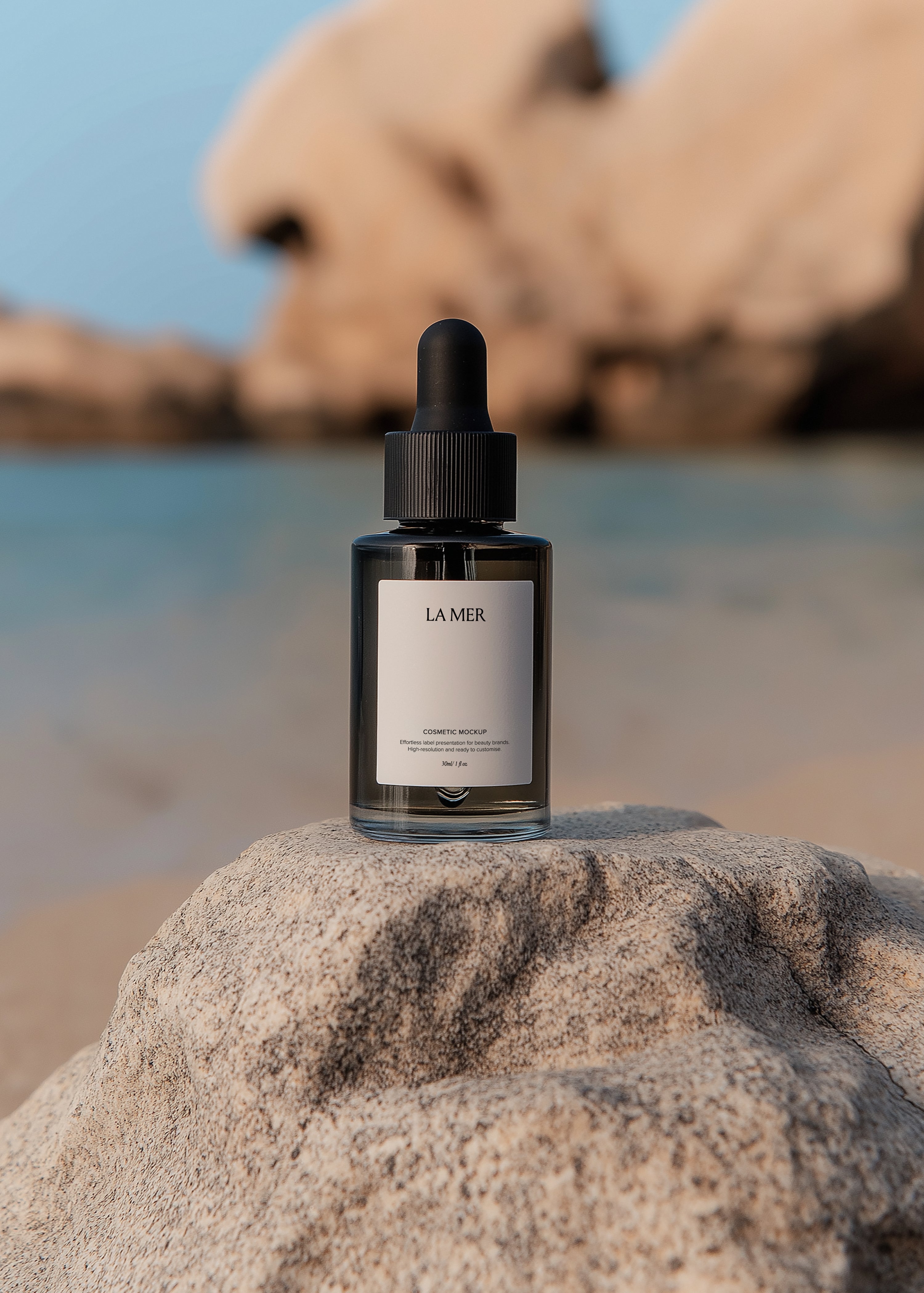 Clear glass dropper bottle with black cap on granite rock, blurred coastal shoreline and boulders in background