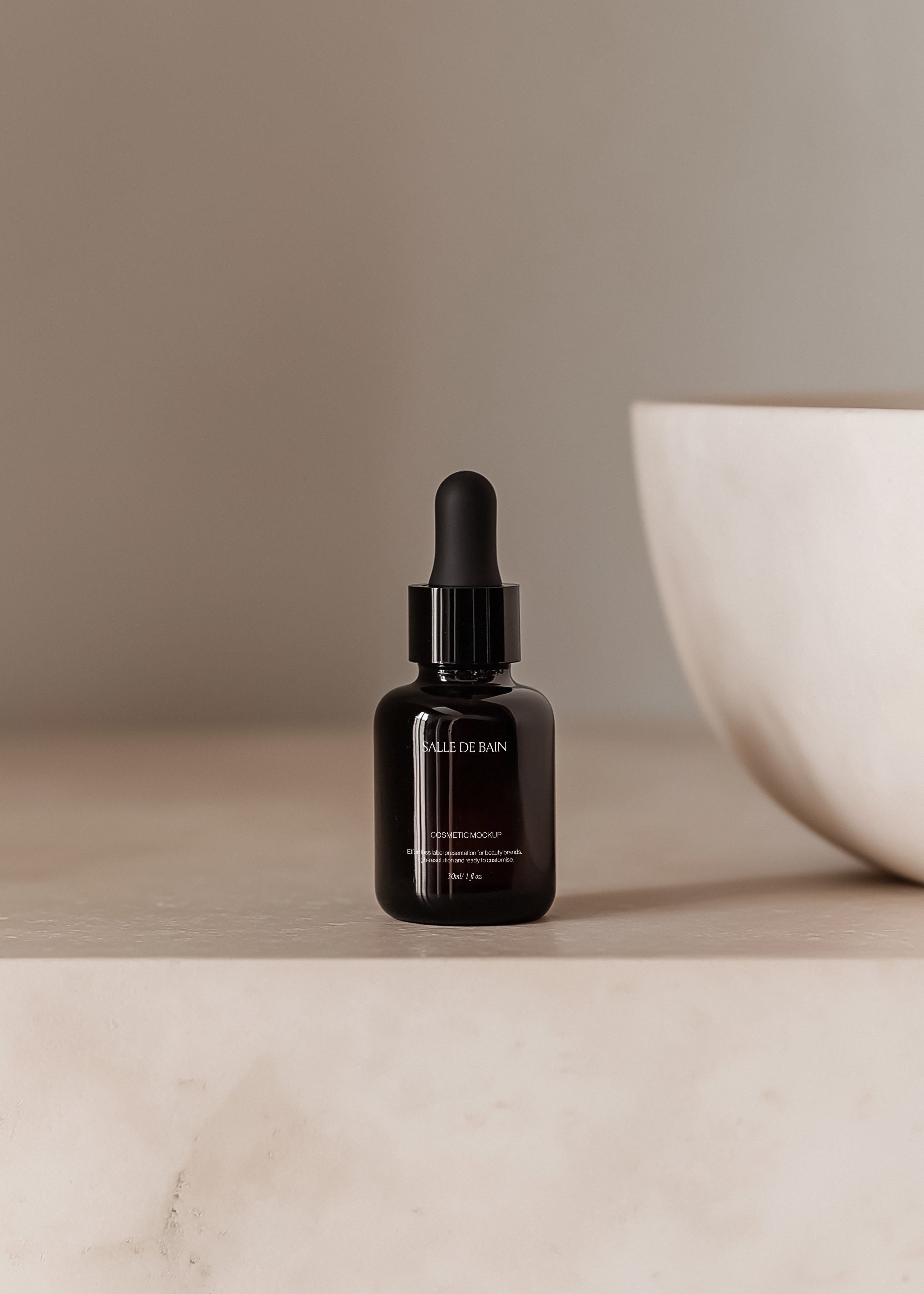 Dark glass dropper bottle mockup standing on pale plaster ledge beside cream ceramic bowl, minimal warm neutral scene