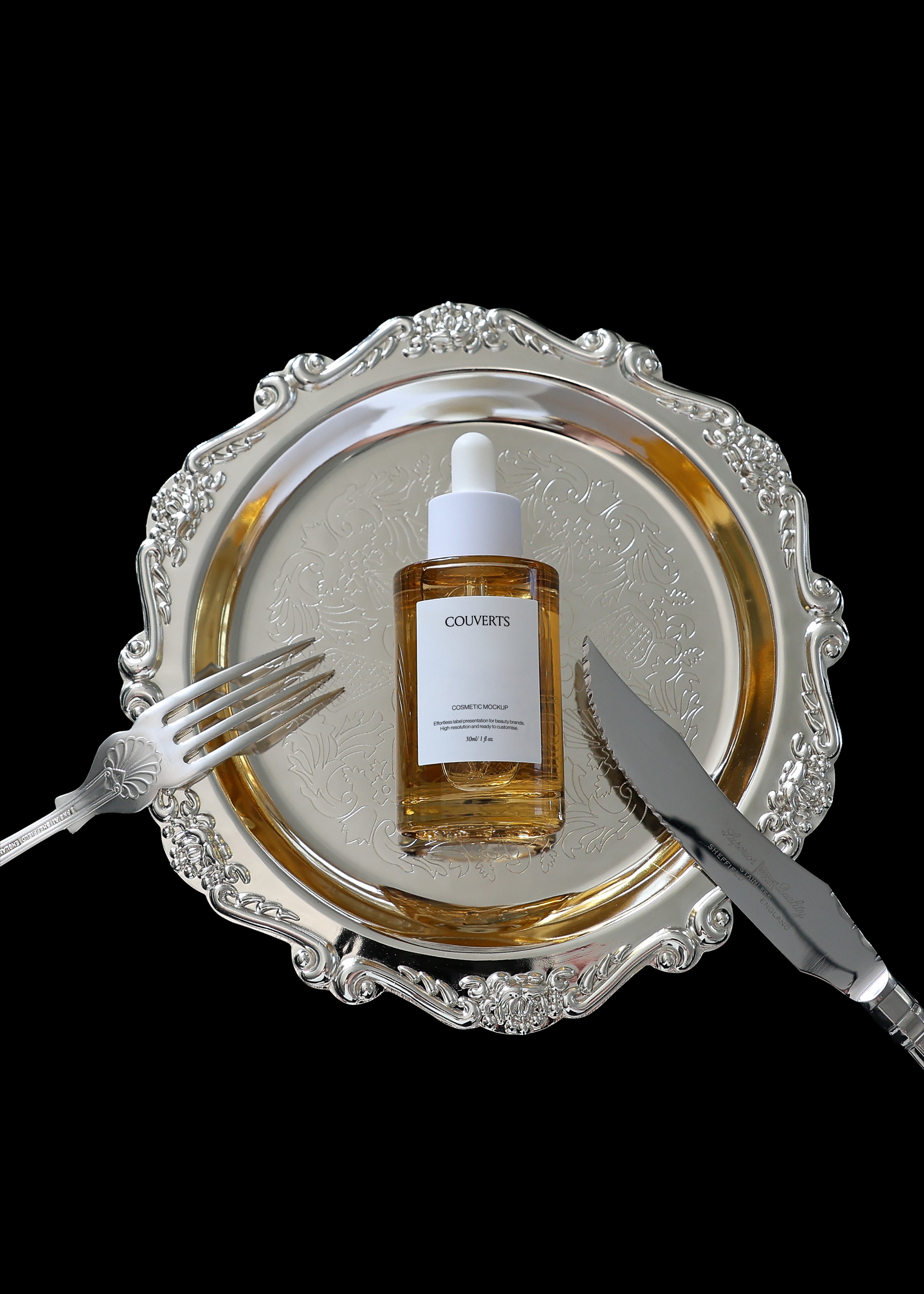 Amber glass dropper bottle mockup on an ornate silver plate with fork and knife, black background, top-down view