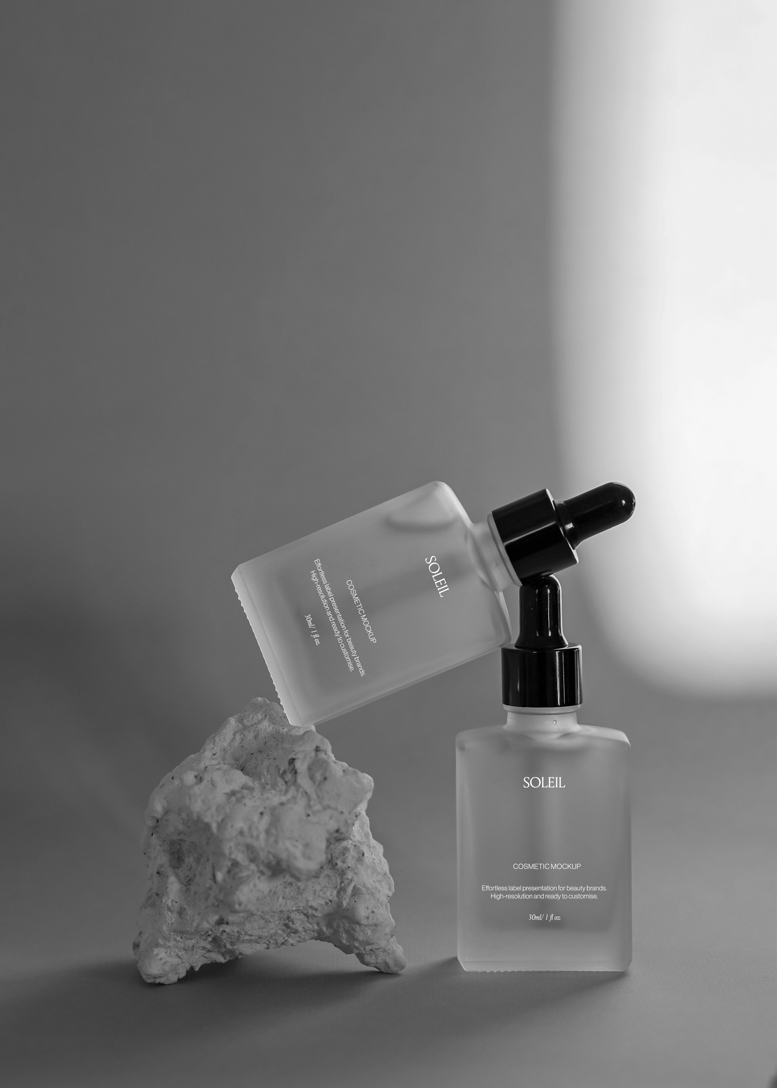 Two frosted glass dropper bottles styled with a rough stone prop, one balanced at an angle, dramatic side lighting on grey backdrop