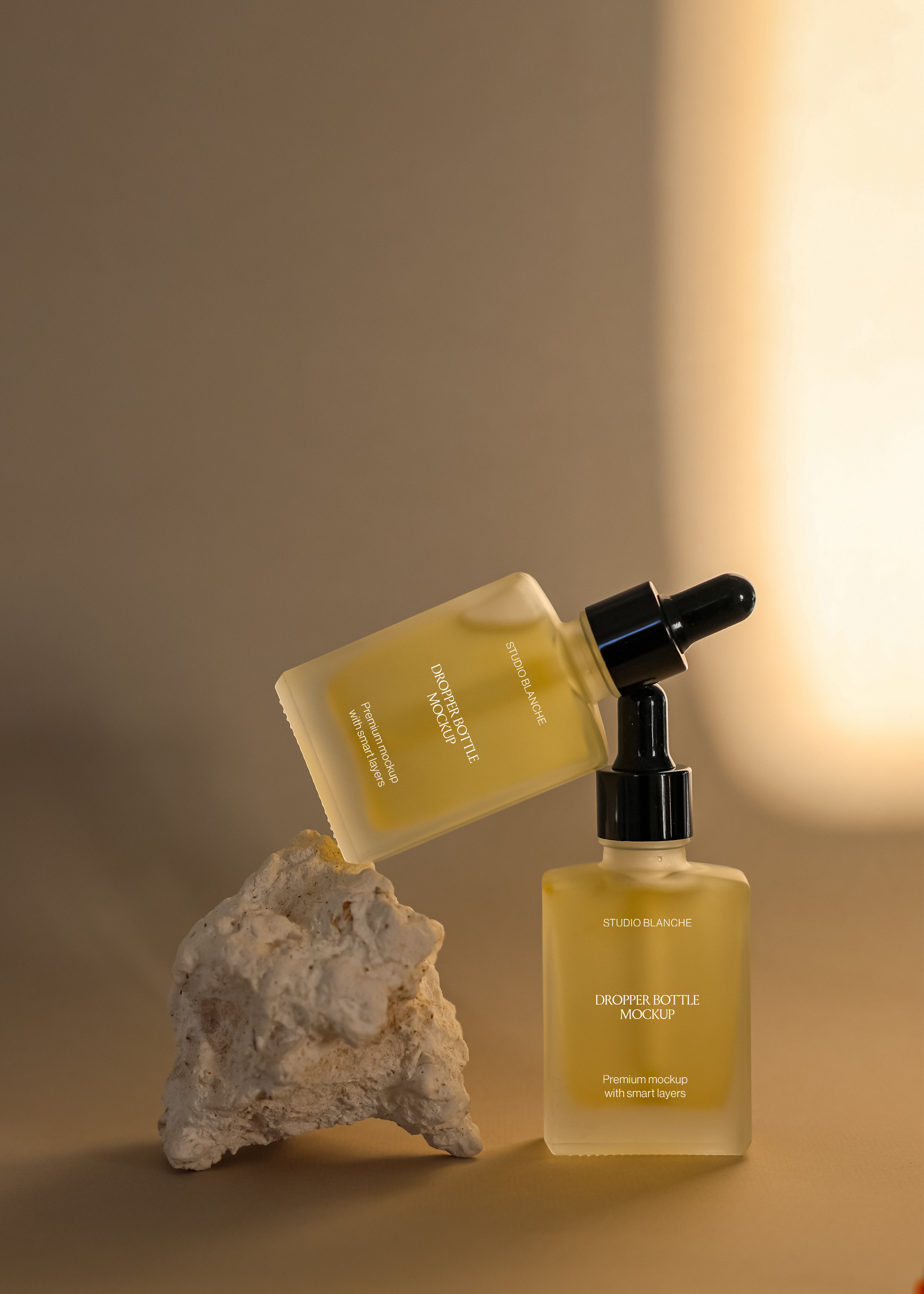 Frosted dropper bottle duo mockup with black pipette caps, warm amber tone version beside stone prop on tan surface