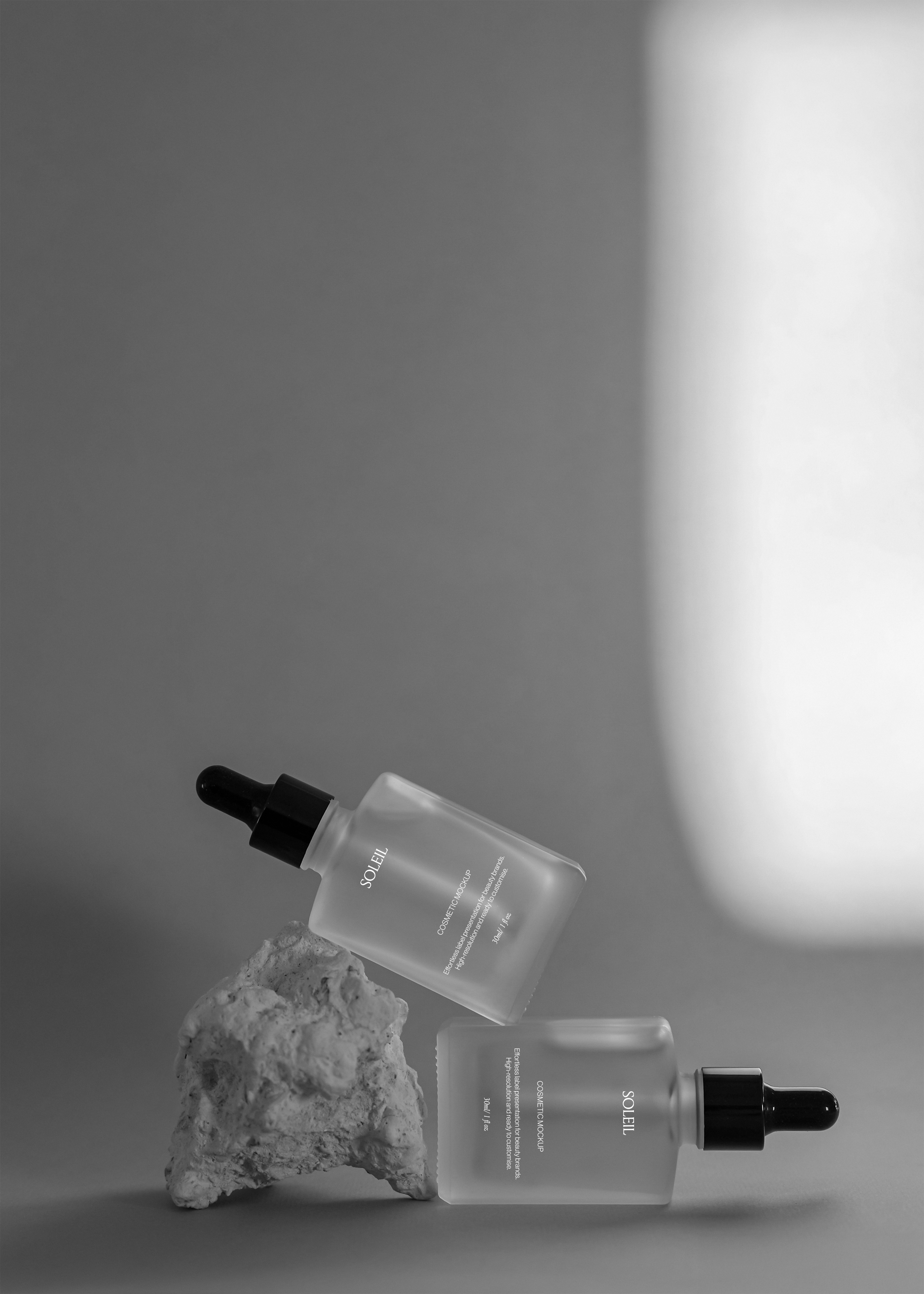 Two frosted glass dropper bottles suspended beside a rough stone prop, wide negative space on grey backdrop, editorial styling