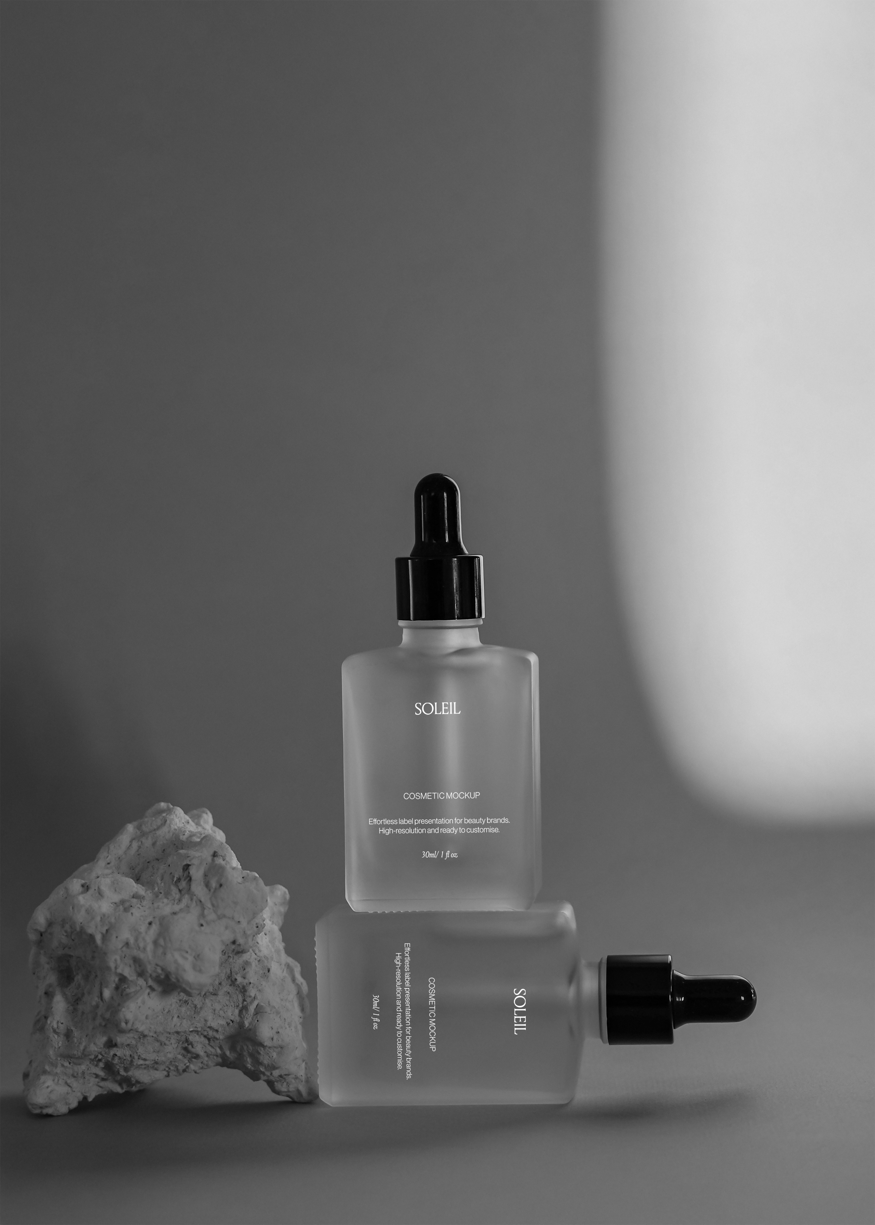 Frosted glass dropper bottle standing upright on a rough stone prop, second bottle lying beside it on grey backdrop