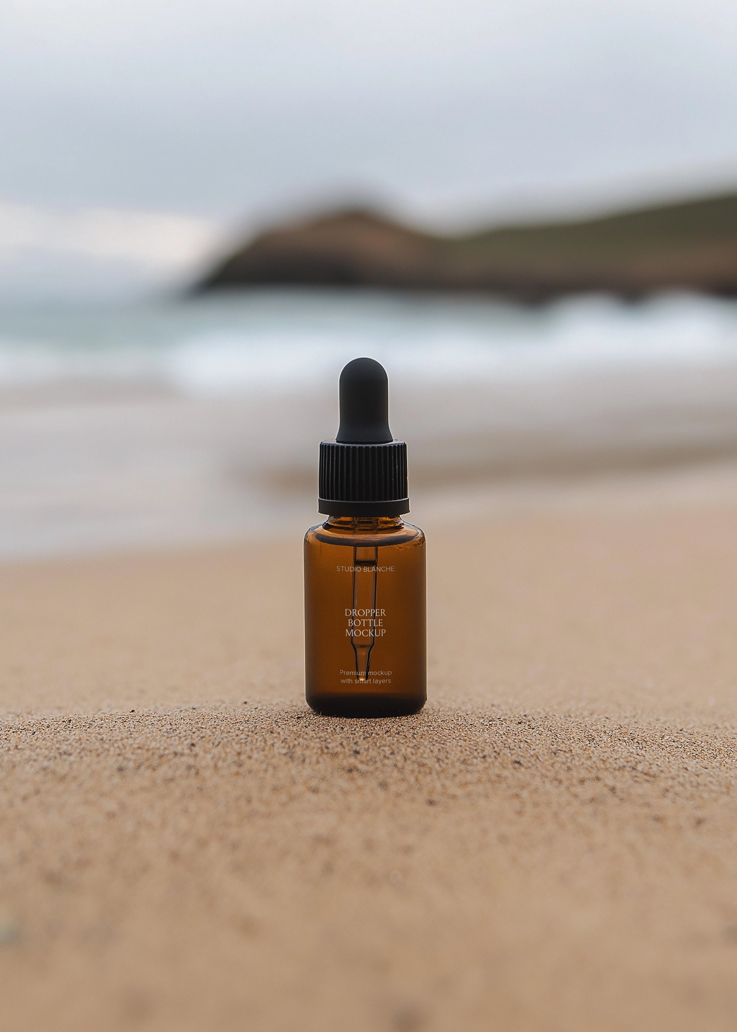 Amber dropper bottle mockup on sandy beach surface, blurred breaking waves and dark rocky headland in soft background