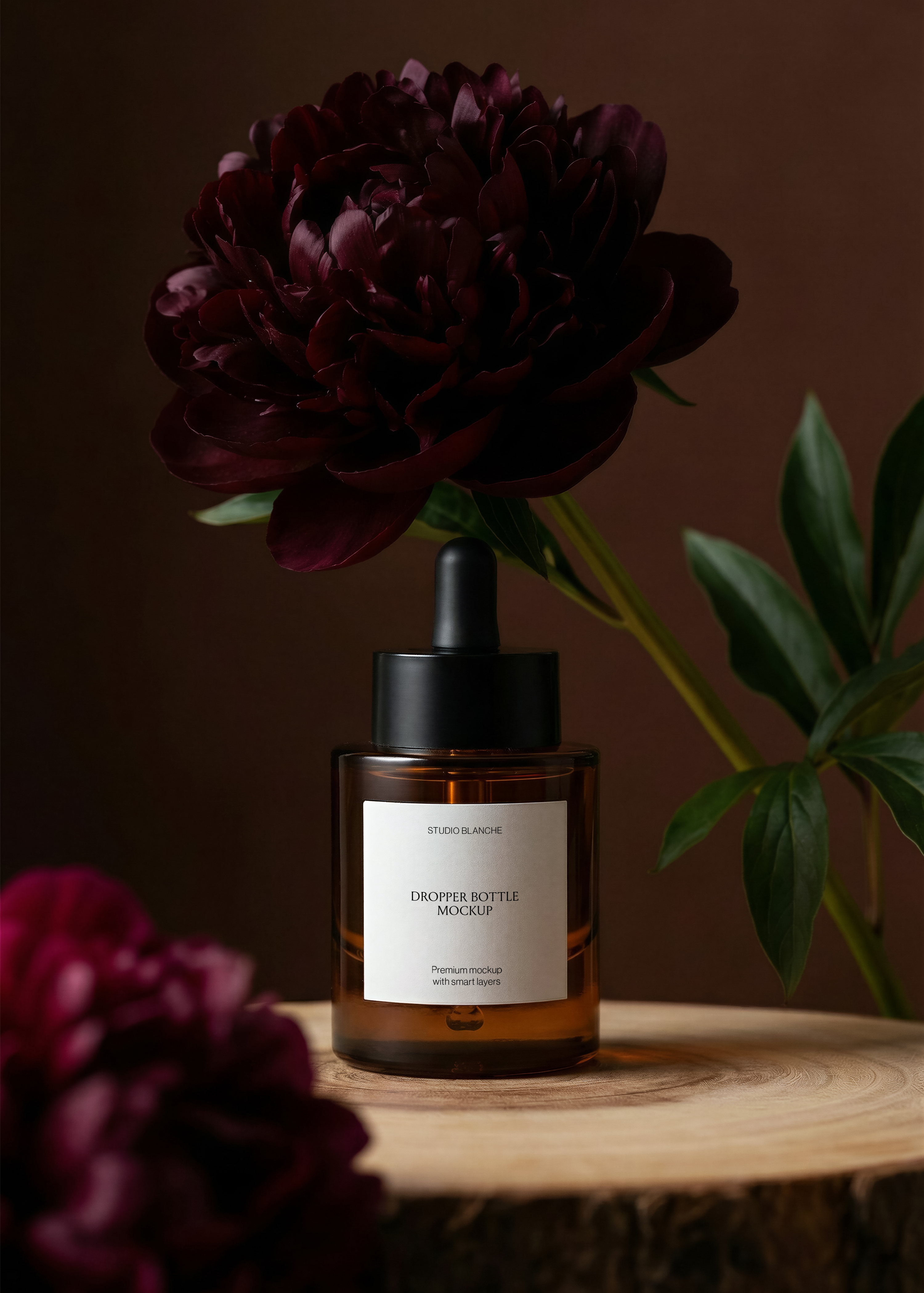Amber dropper bottle mockup on wood slice, dark burgundy peony overhead, green foliage, blurred foreground bloom, dark background
