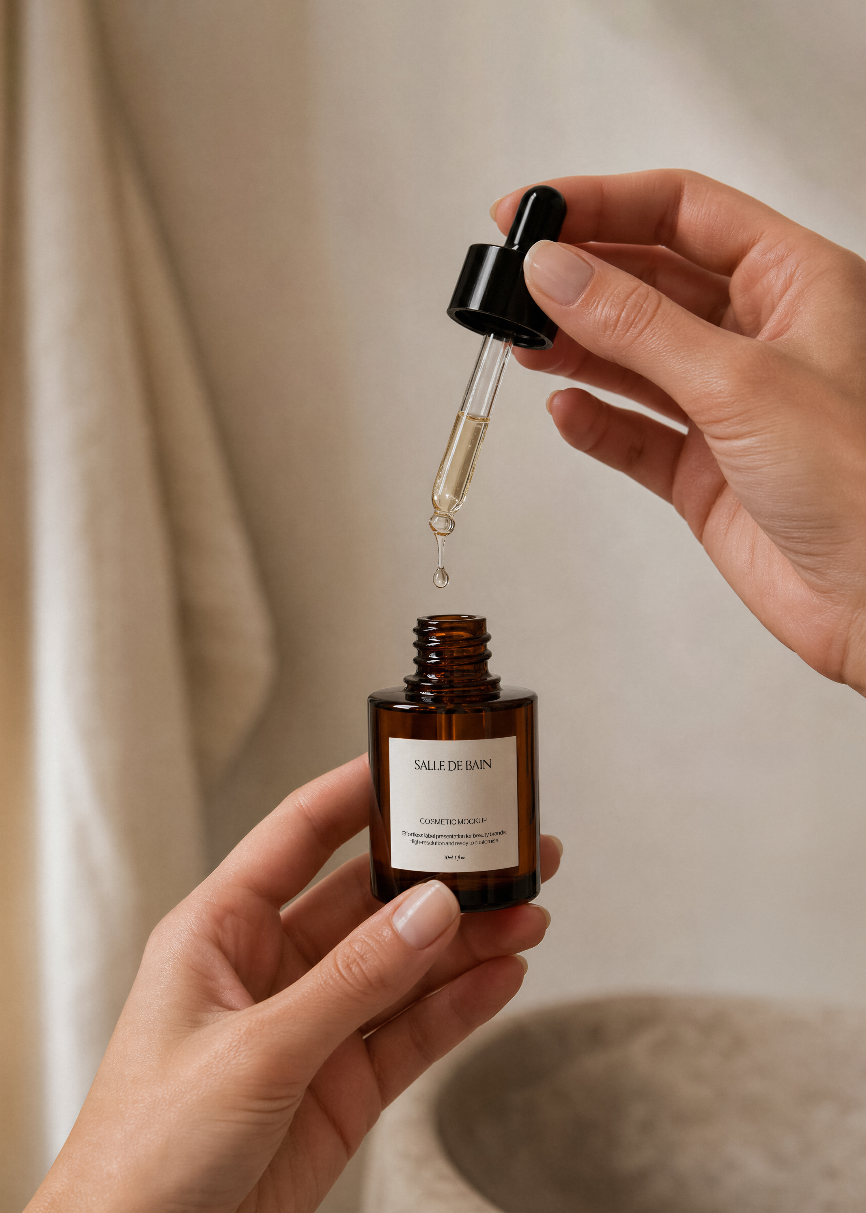 Amber glass dropper bottle mockup held in two hands with dropper dispensing an oil drop, warm neutral backdrop