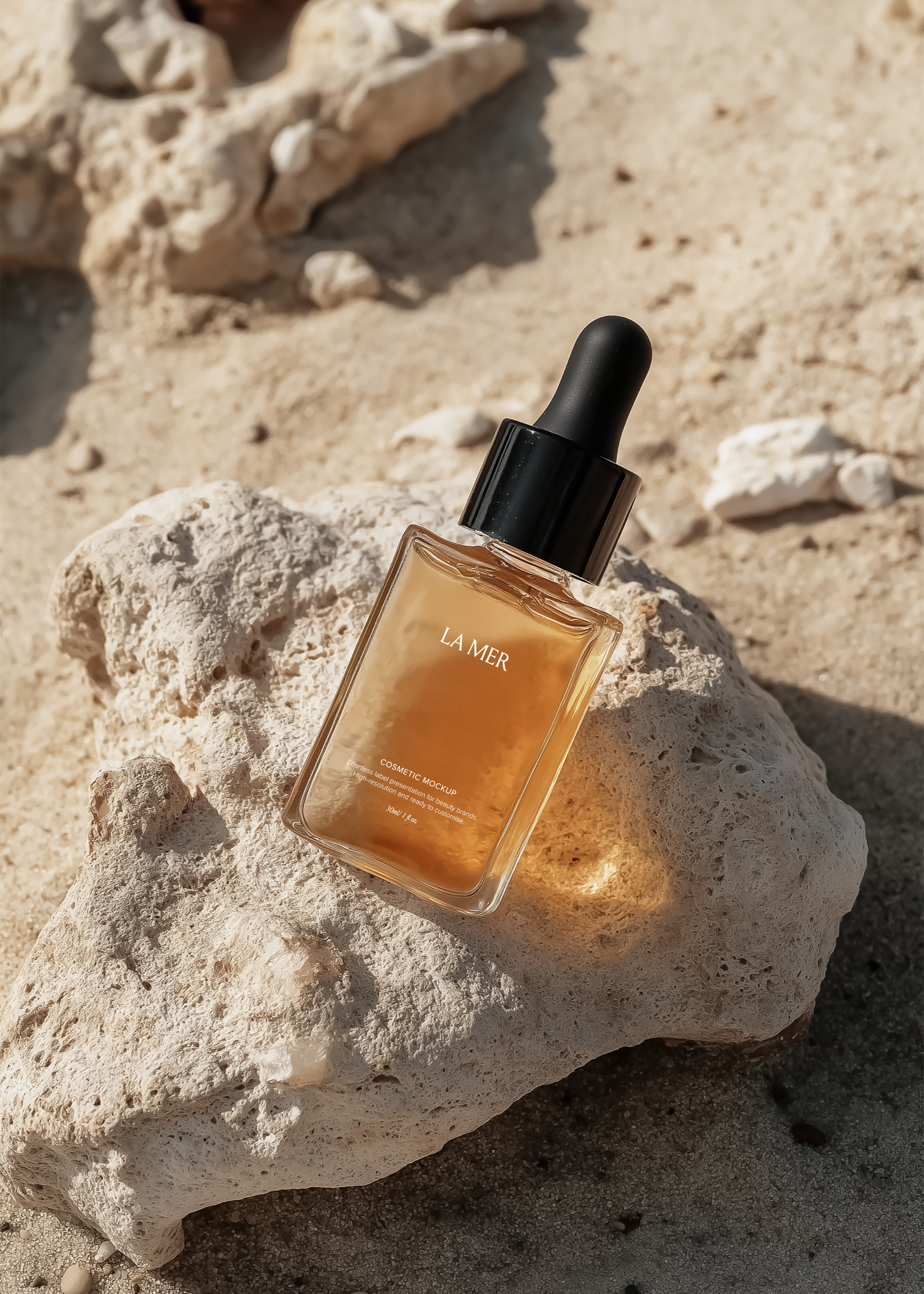 Gold-toned glass dropper bottle with black cap resting on bleached coral rock, warm sand and sunlight, angled view
