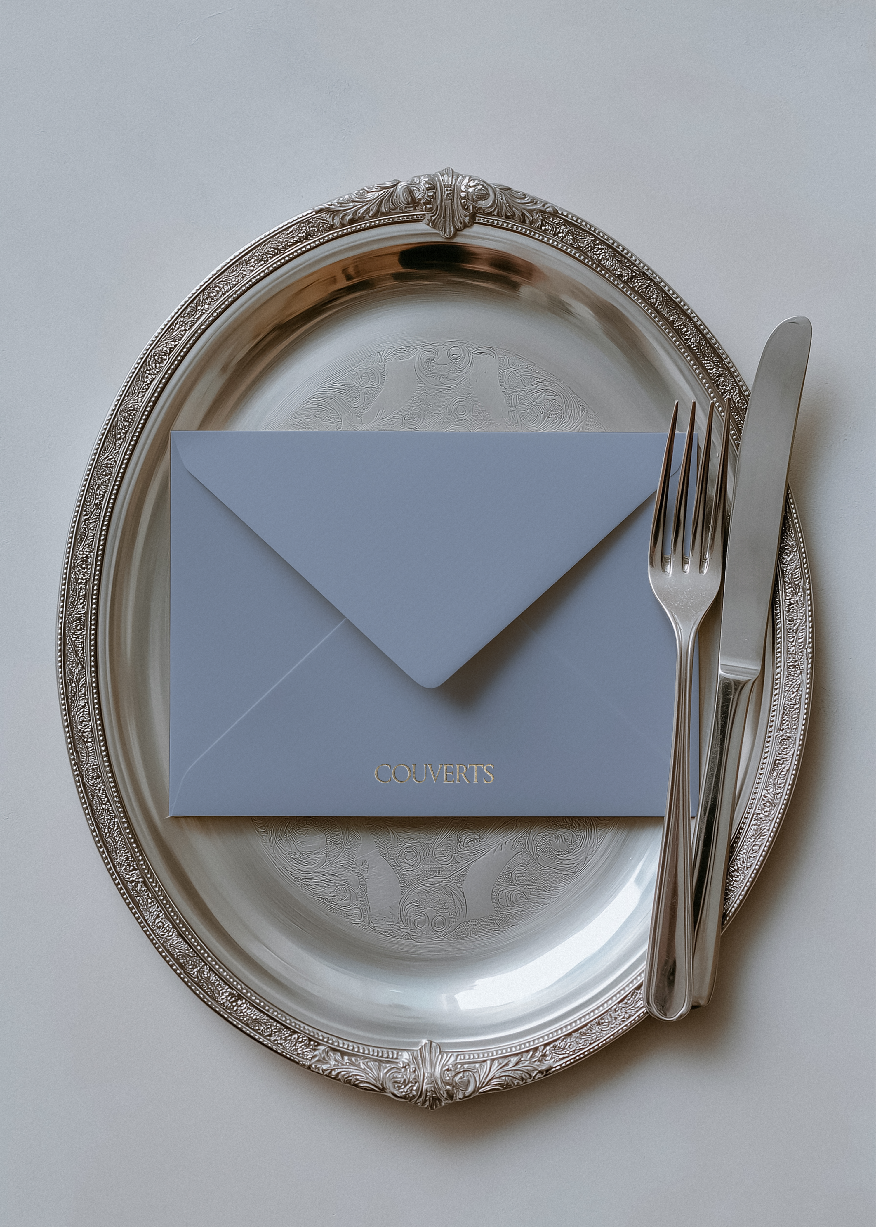 Dove blue envelope mockup with gold foil text, set on ornate silver charger beside fork and knife, ivory backdrop