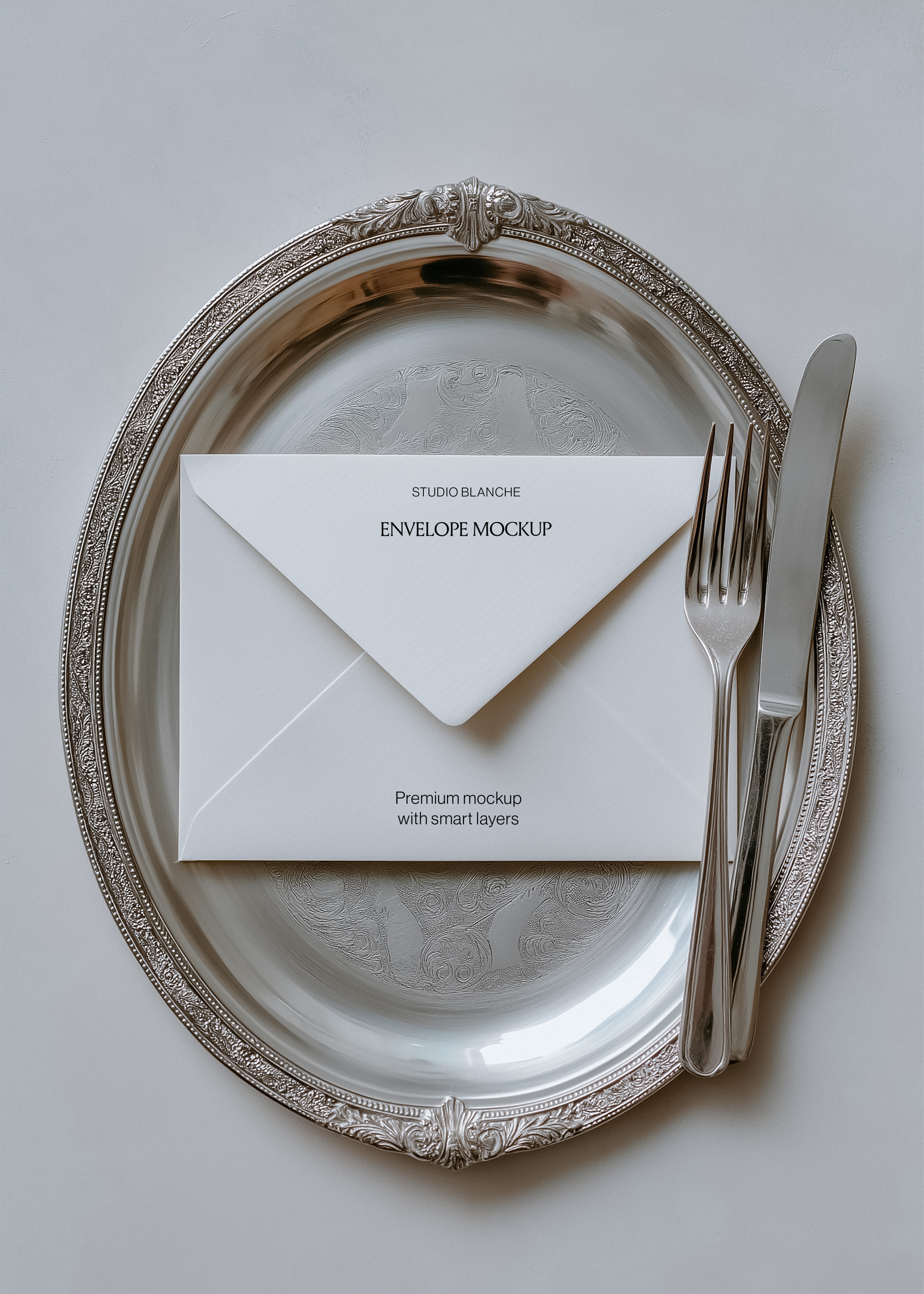 Envelope mockup with Studio Blanche wordmark on cream paper, resting on engraved silver charger with cutlery, soft light styling
