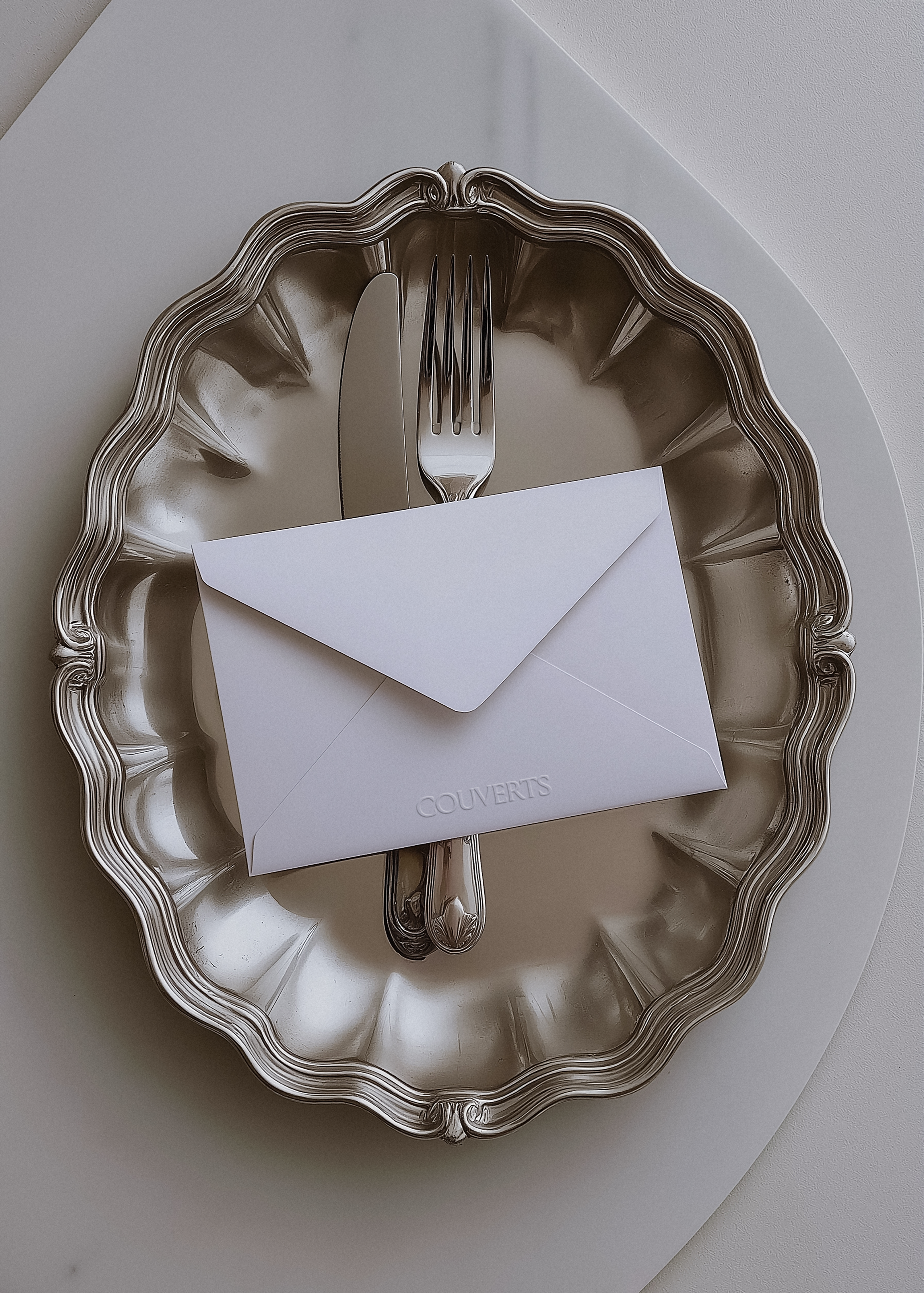 White embossed envelope mockup on scalloped silver tray beside fork and knife, soft grey angled surface