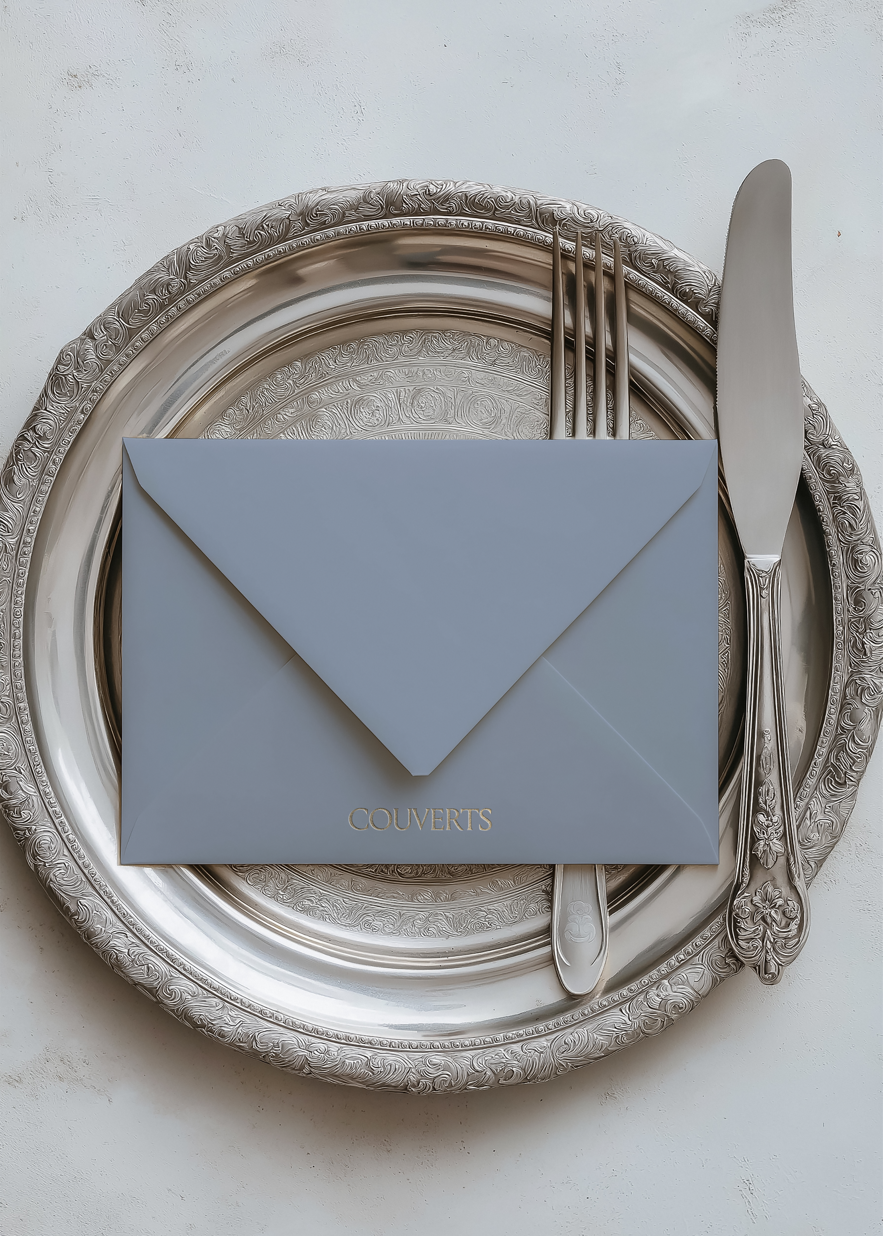 Dove blue envelope mockup with gold foil text on ornate silver tray beside monogrammed fork and knife, plaster backdrop