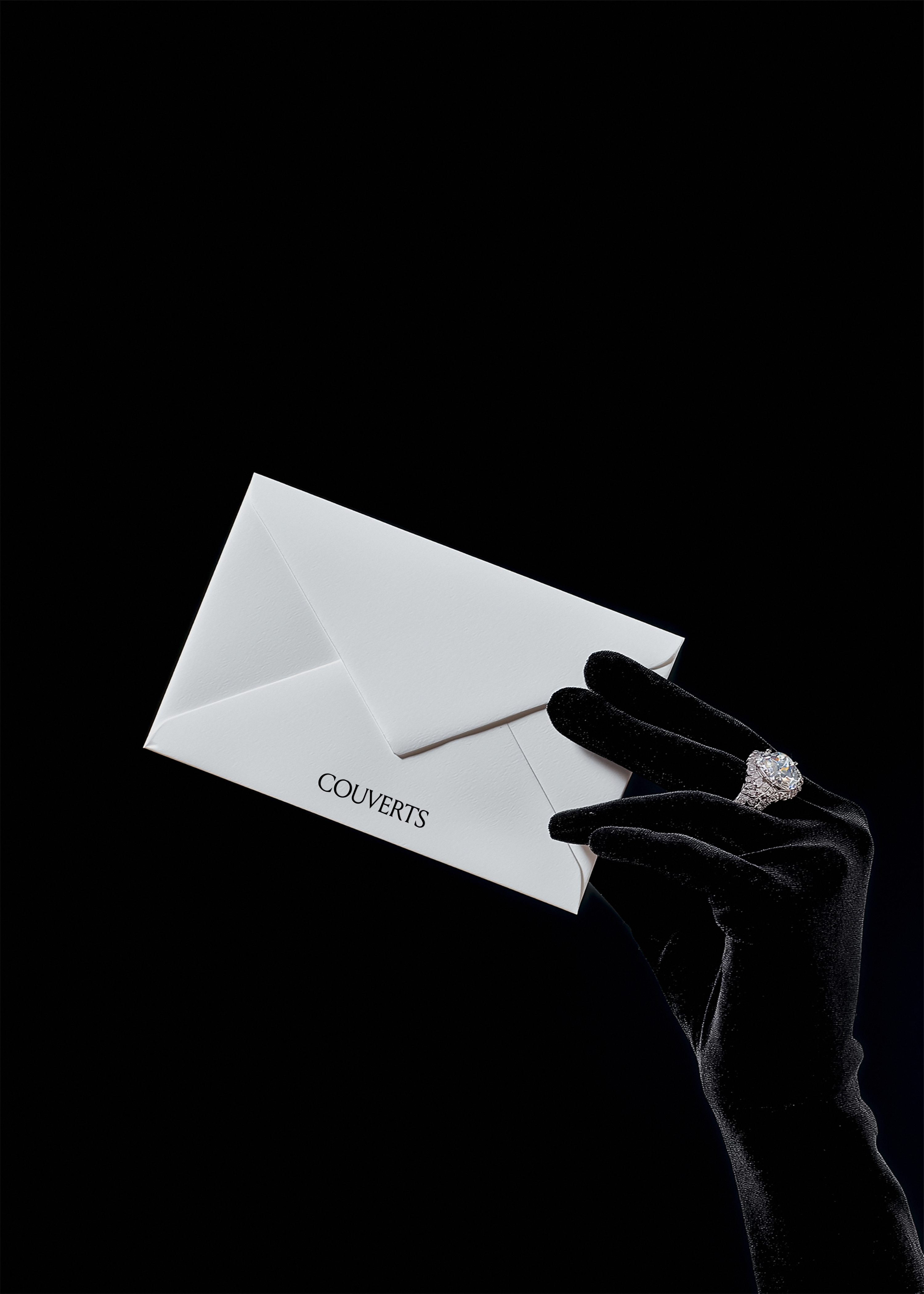 White envelope mockup tilted mid air, held loosely by black satin gloved fingers with diamond ring, black backdrop