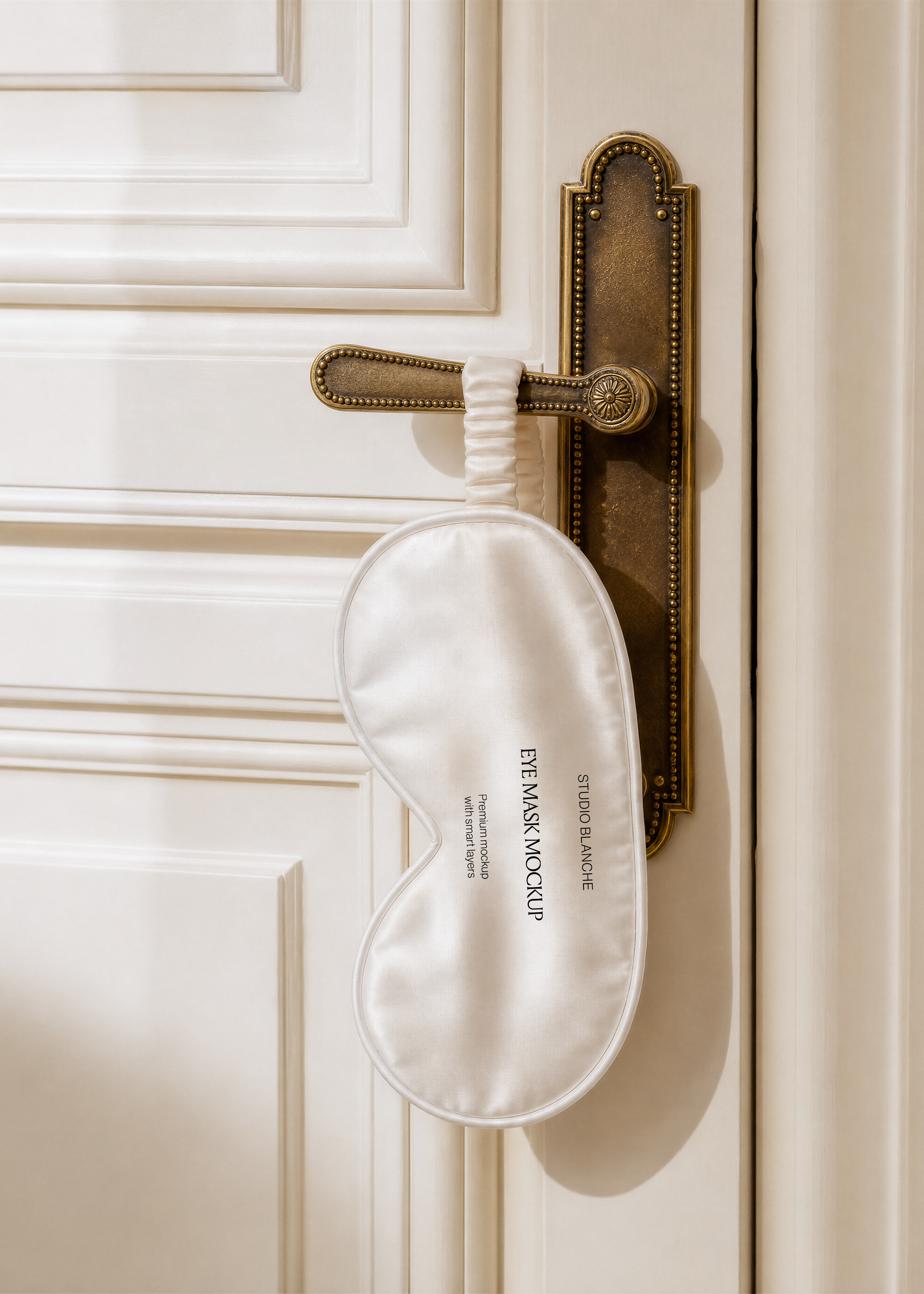 Cream silk eye mask mockup with elasticated strap looped over a brass handle plate on a panelled door