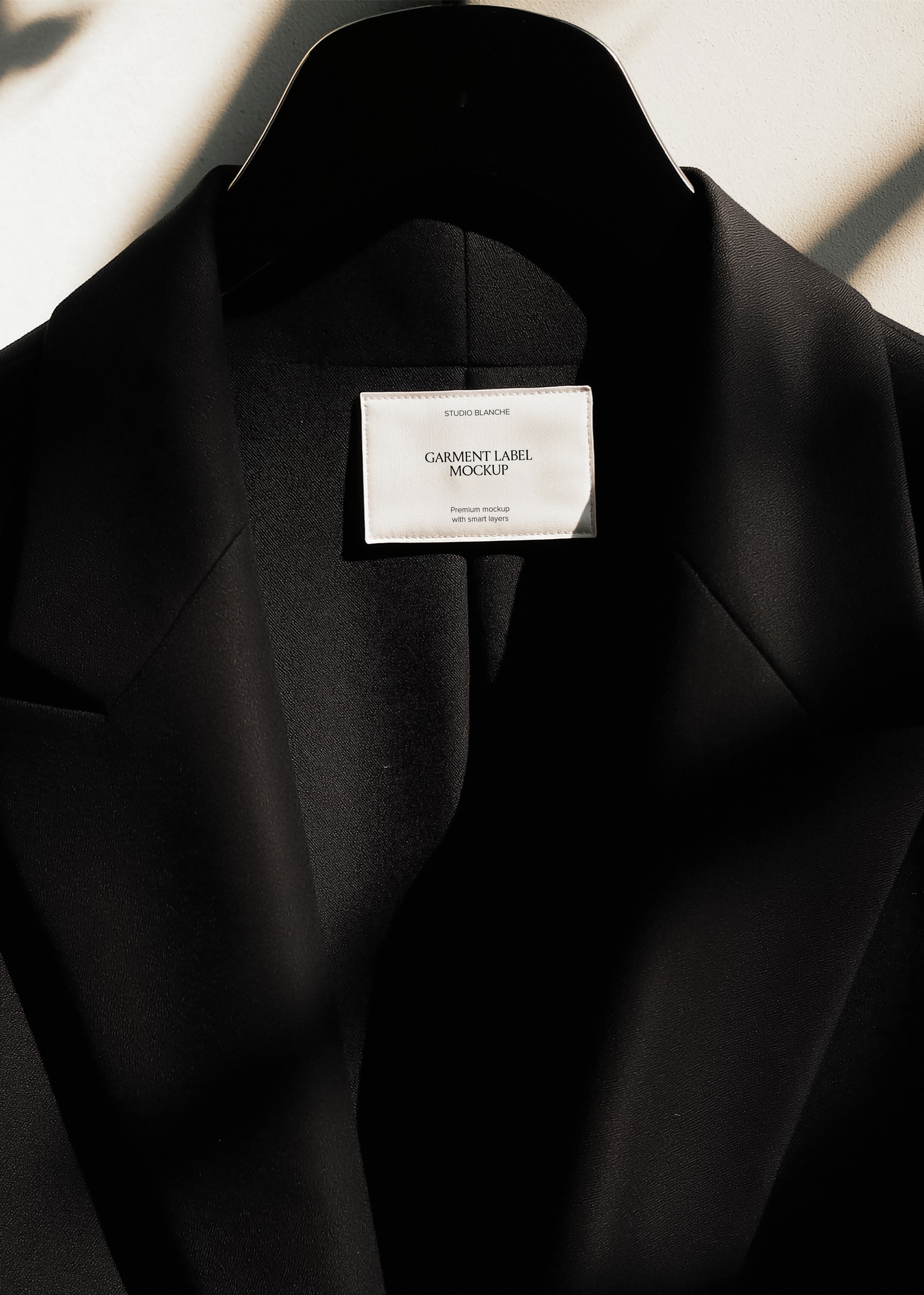 White garment label mockup inside collar of black blazer on hanger, dappled natural light against cream wall 