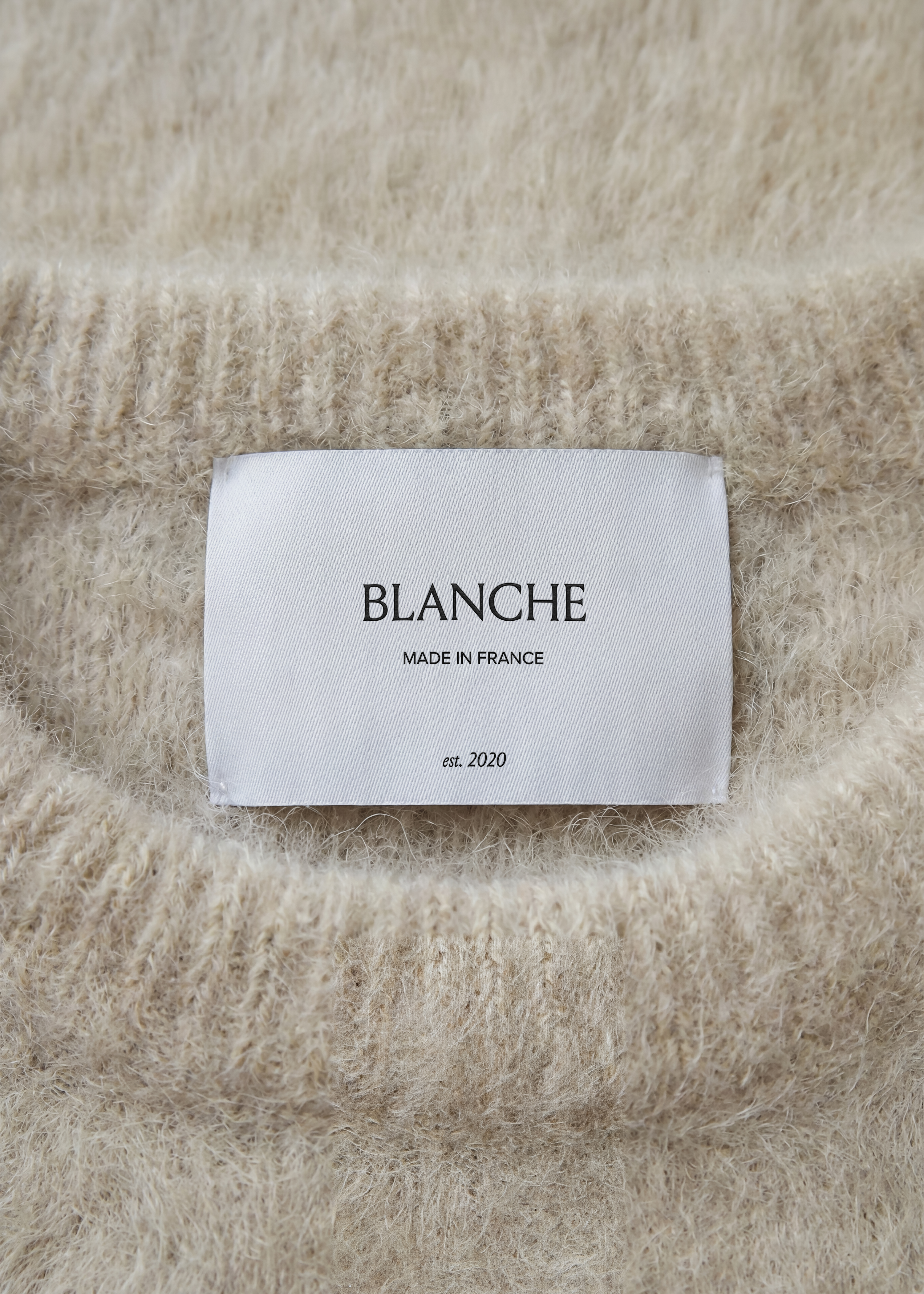 Satin garment label mockup centred on a brushed sand mohair-style knit, soft tactile fashion close-up in pale tones