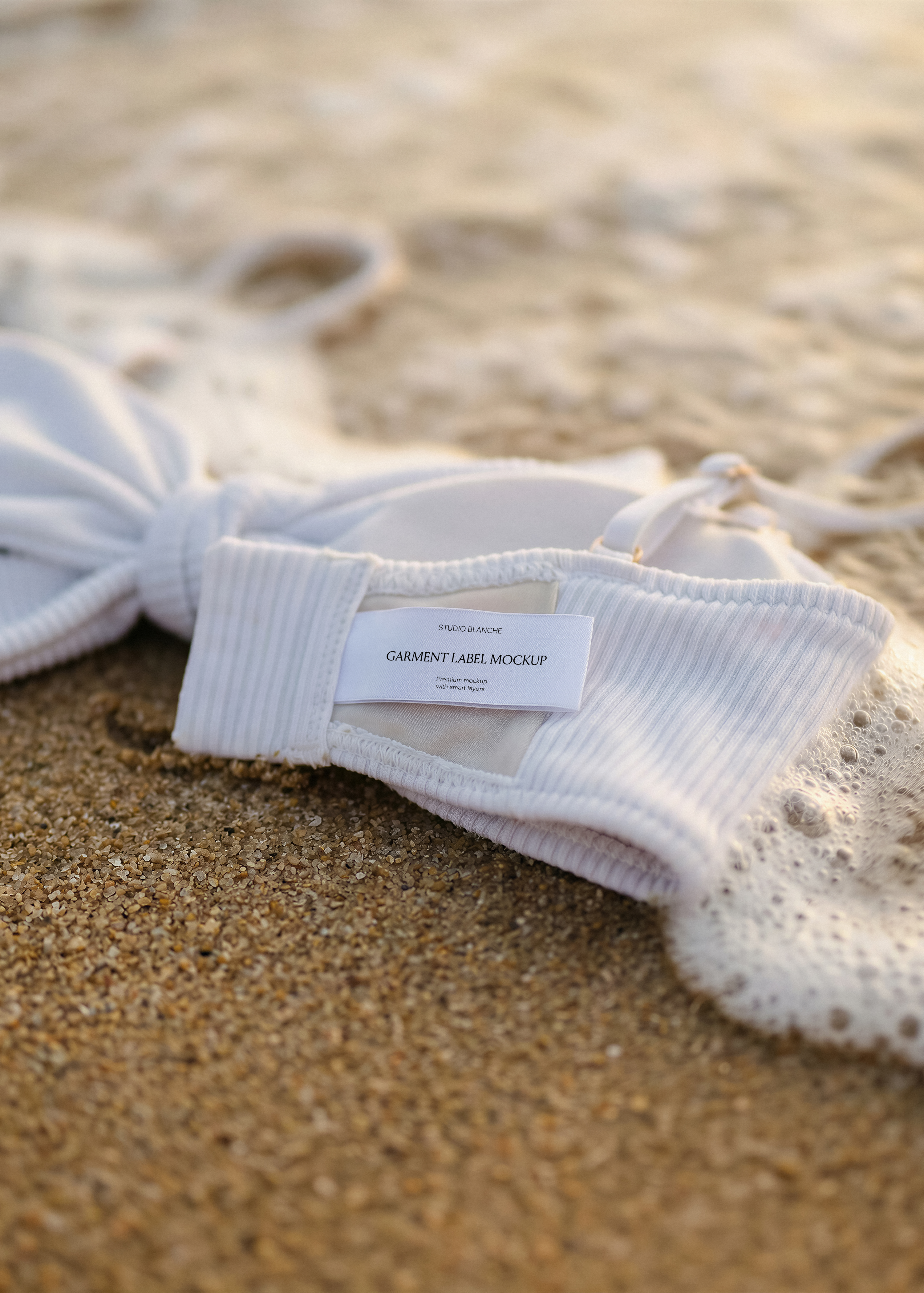 White woven label on white ribbed bikini top resting on wet sand at the shoreline, coastal lifestyle scene 