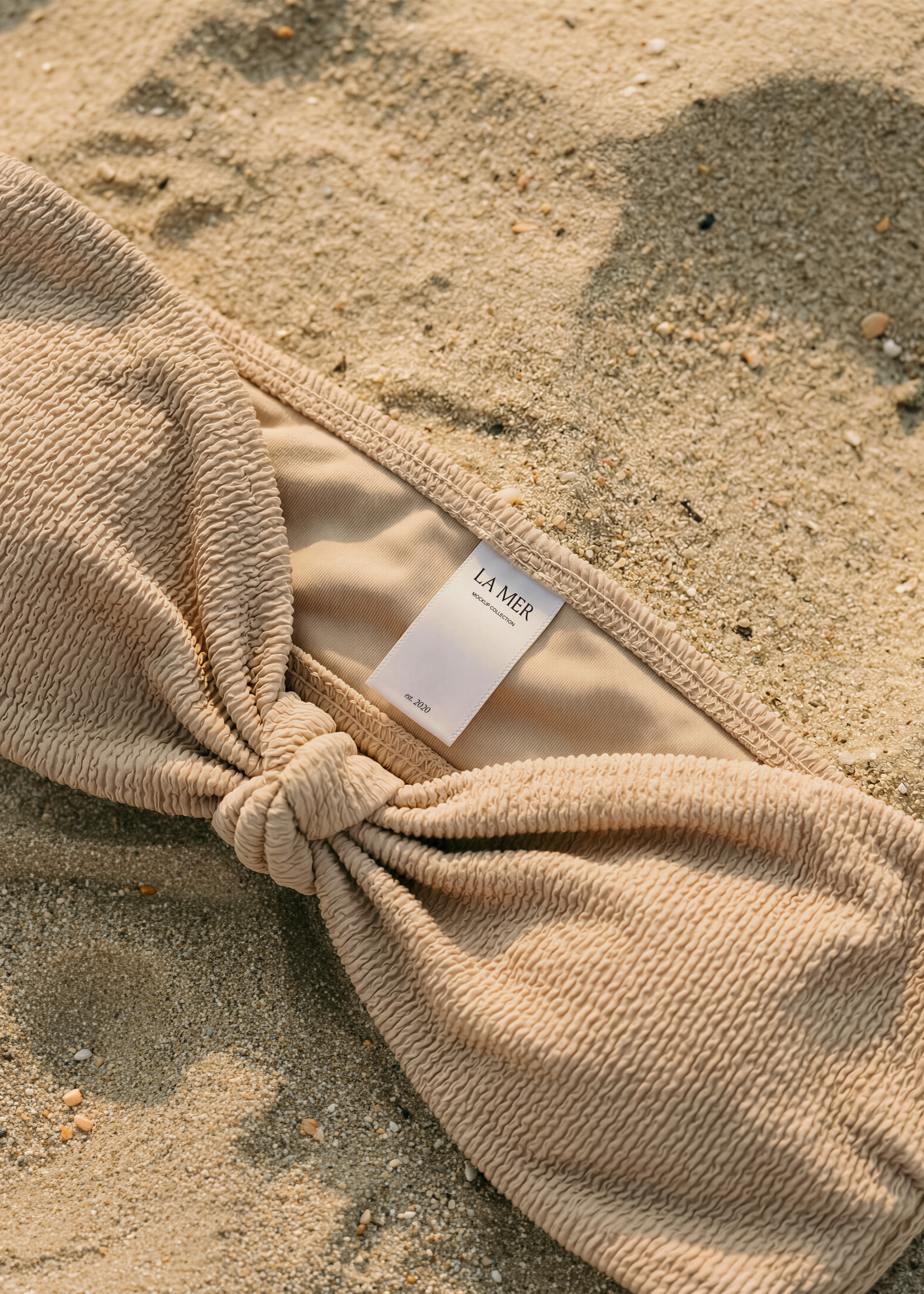 Garment label mockup on sand-tone crinkle bikini top, inner lining visible, dry sand background with shells