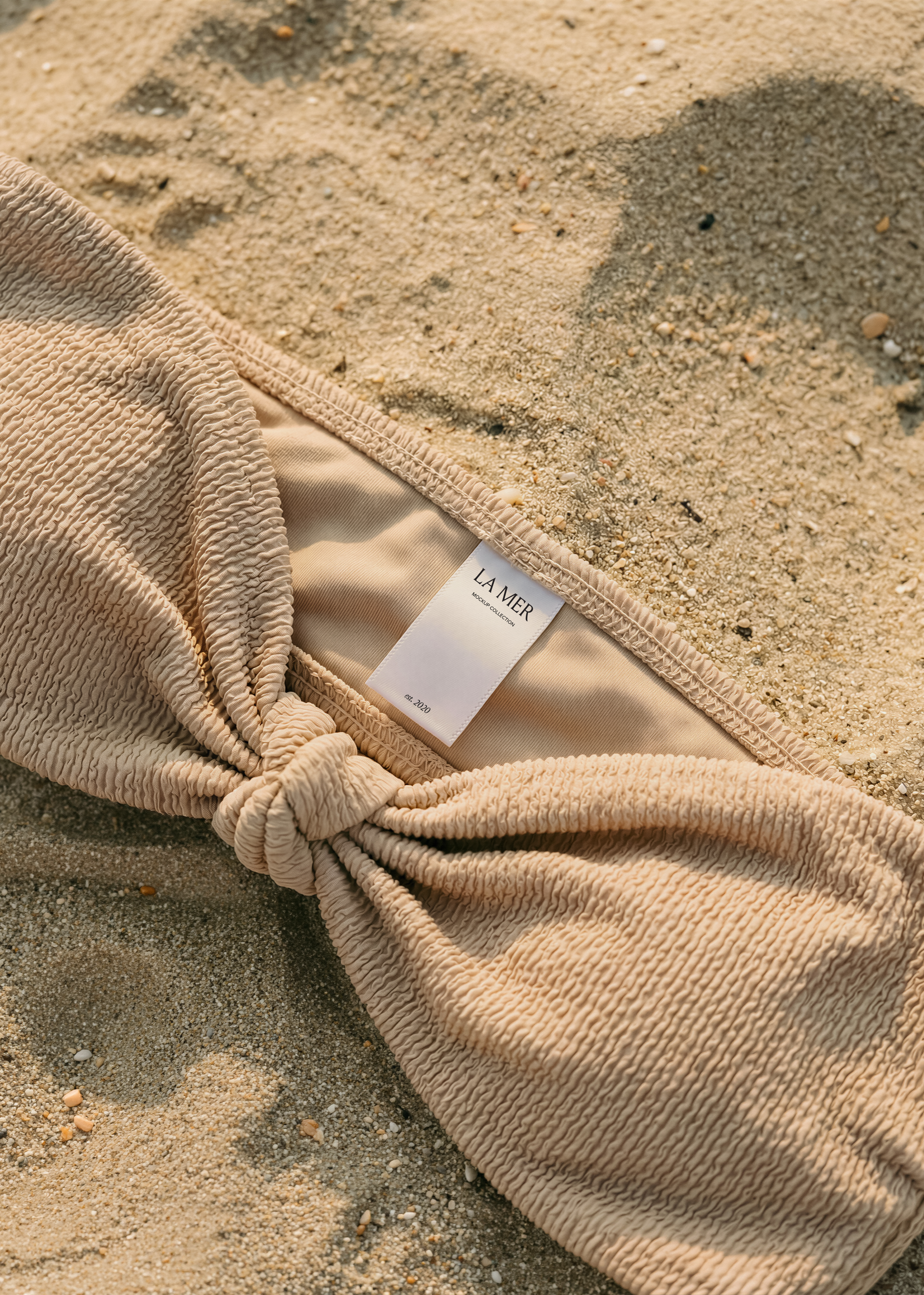 Garment label mockup on sand-tone crinkle bikini top, inner lining visible, dry sand background with shells