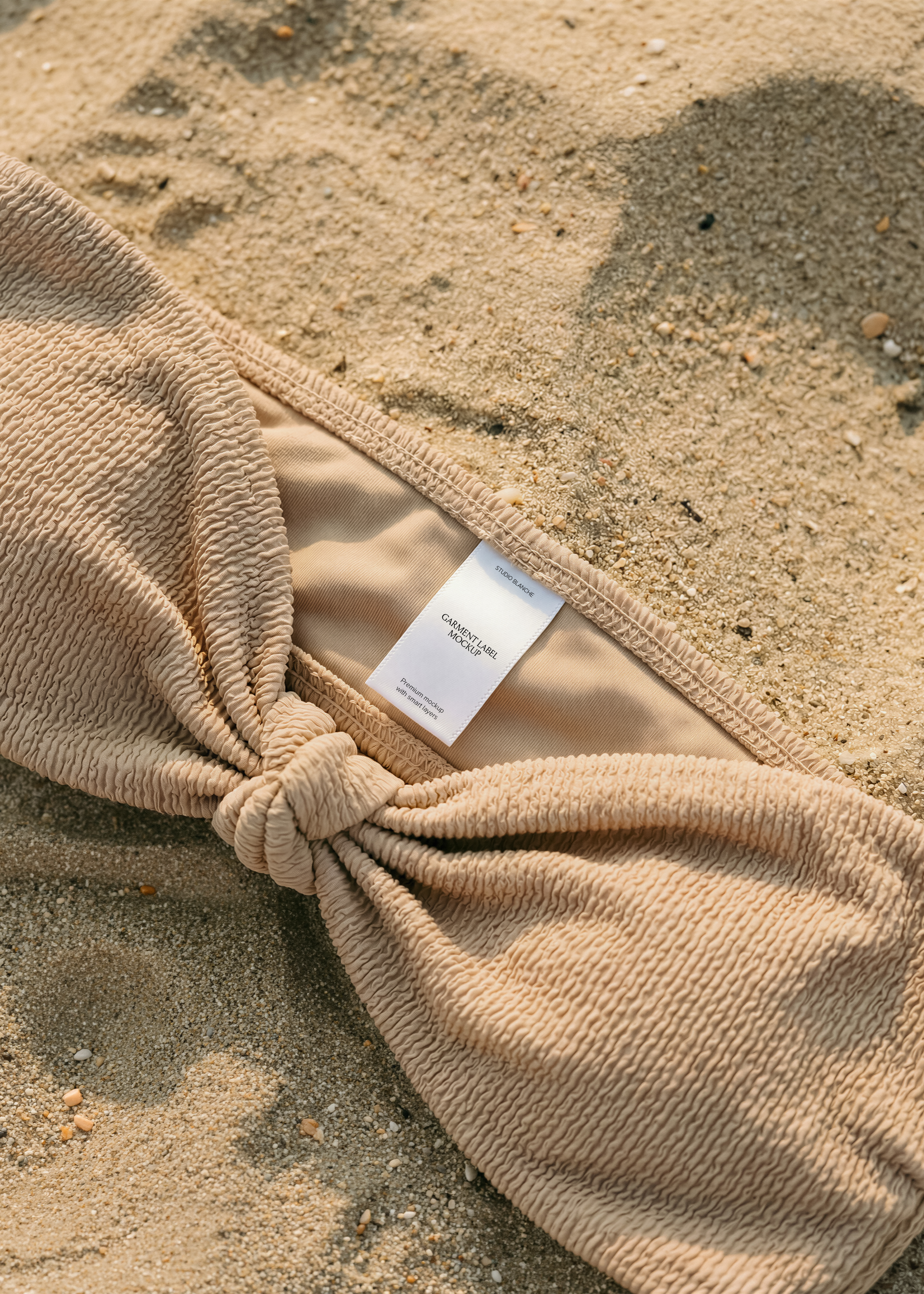 White woven label inside taupe crinkle bandeau bikini top laid flat on dry beach sand, warm tonal styling