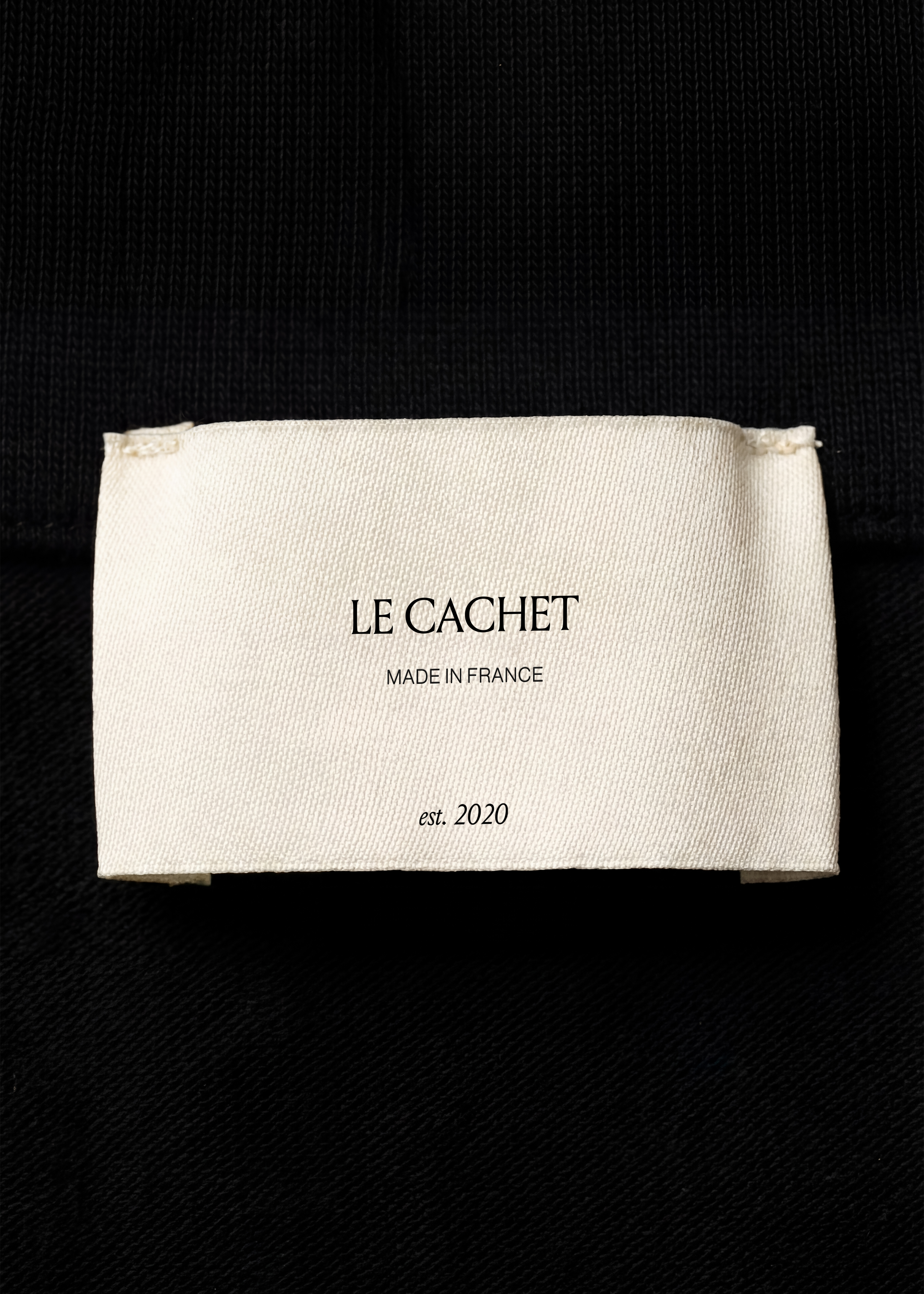 Cream cotton garment label mockup stitched onto black ribbed and twill fabric, close-up macro detail, monochrome styling