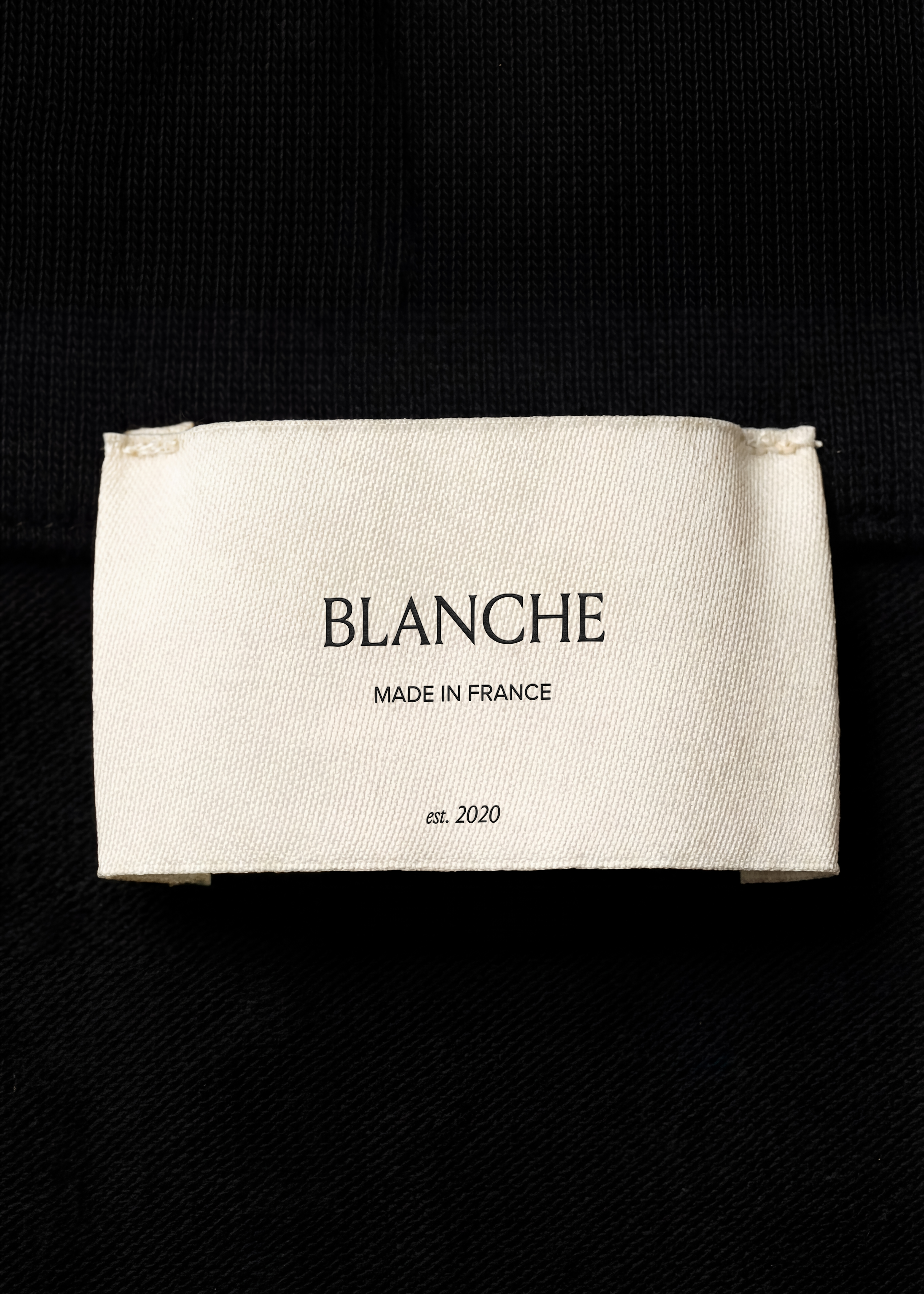 Cream cotton garment label mockup stitched onto black ribbed and twill fabric, close-up macro detail, monochrome styling