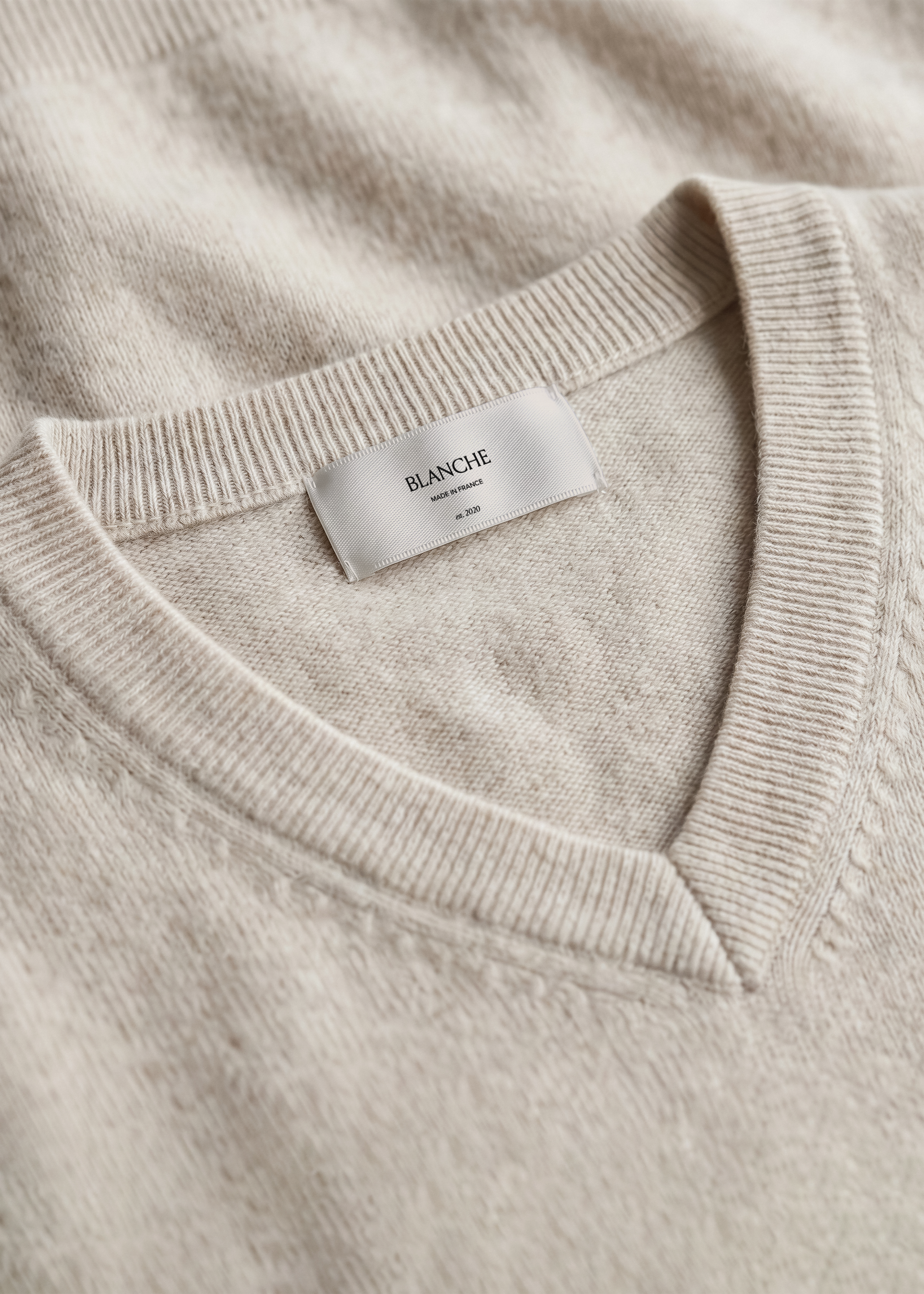 Satin garment label mockup stitched into the v-neckline of an oatmeal cashmere jumper, soft tonal fashion close-up