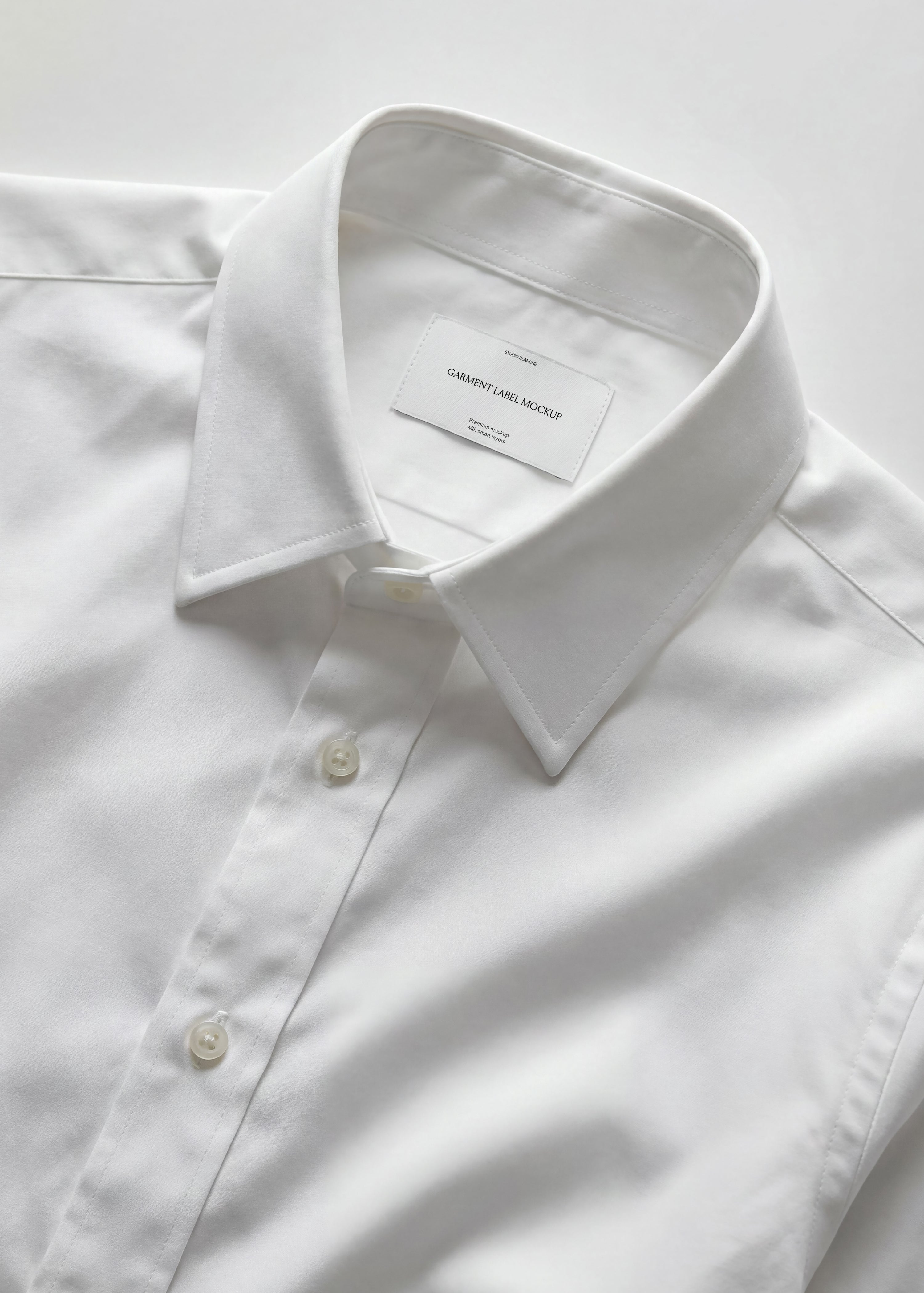 Woven garment label mockup on white cotton shirt collar with pearl buttons, crisp tailoring detail in tonal whites