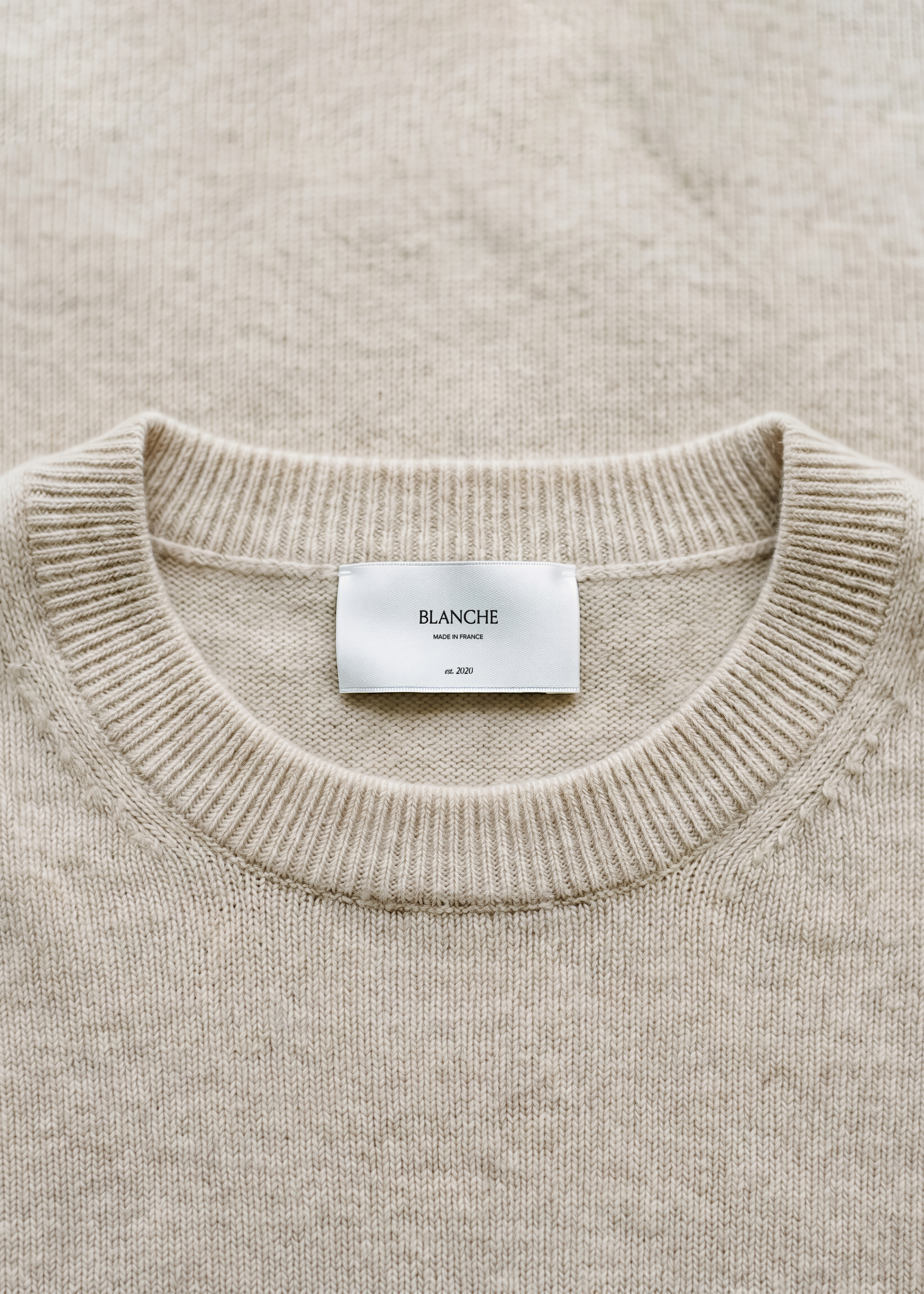 Satin garment label mockup centred on the ribbed crew neckline of an oatmeal cashmere jumper, soft tonal flat-lay