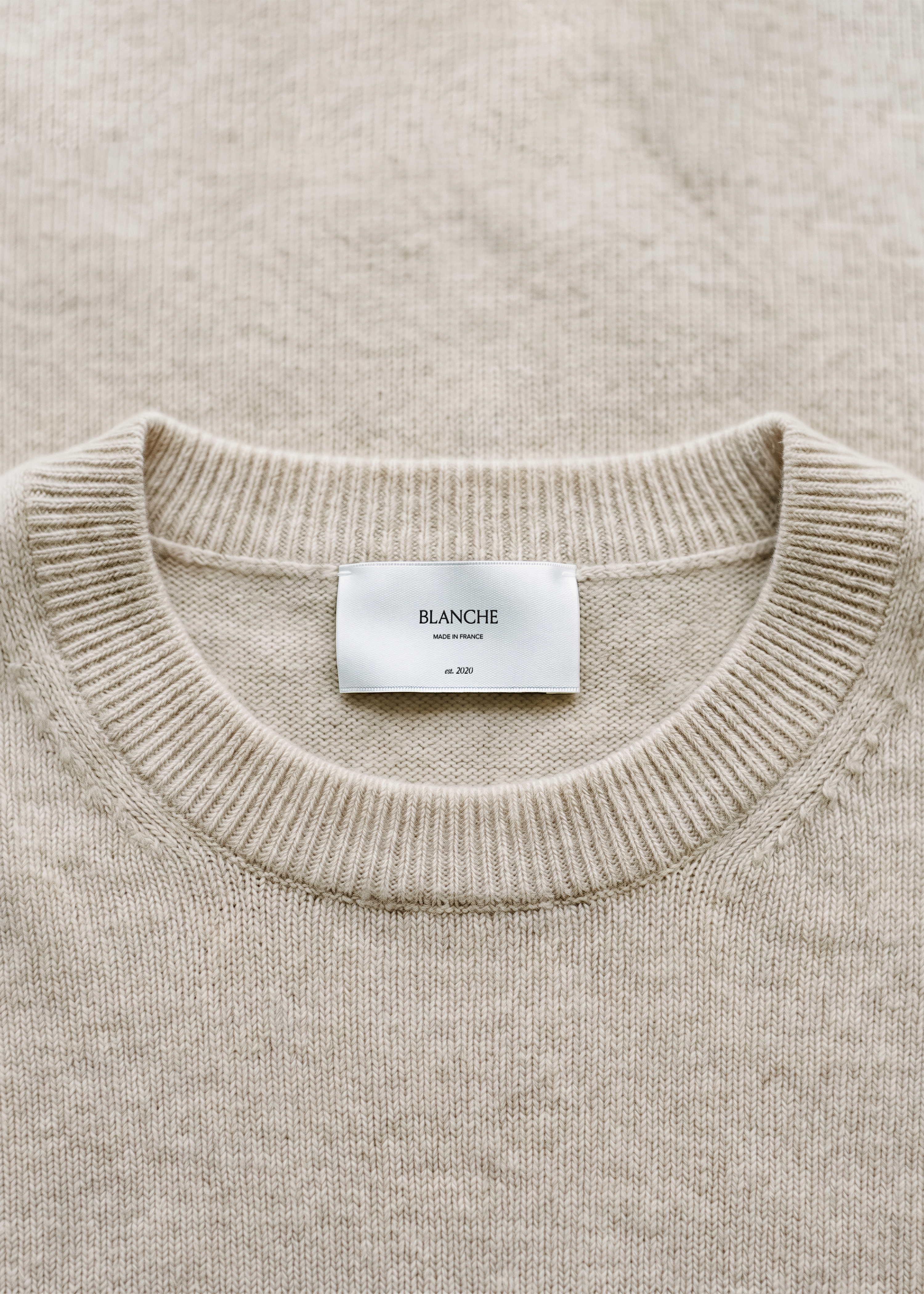 Satin garment label mockup centred on the ribbed crew neckline of an oatmeal cashmere jumper, soft tonal flat-lay