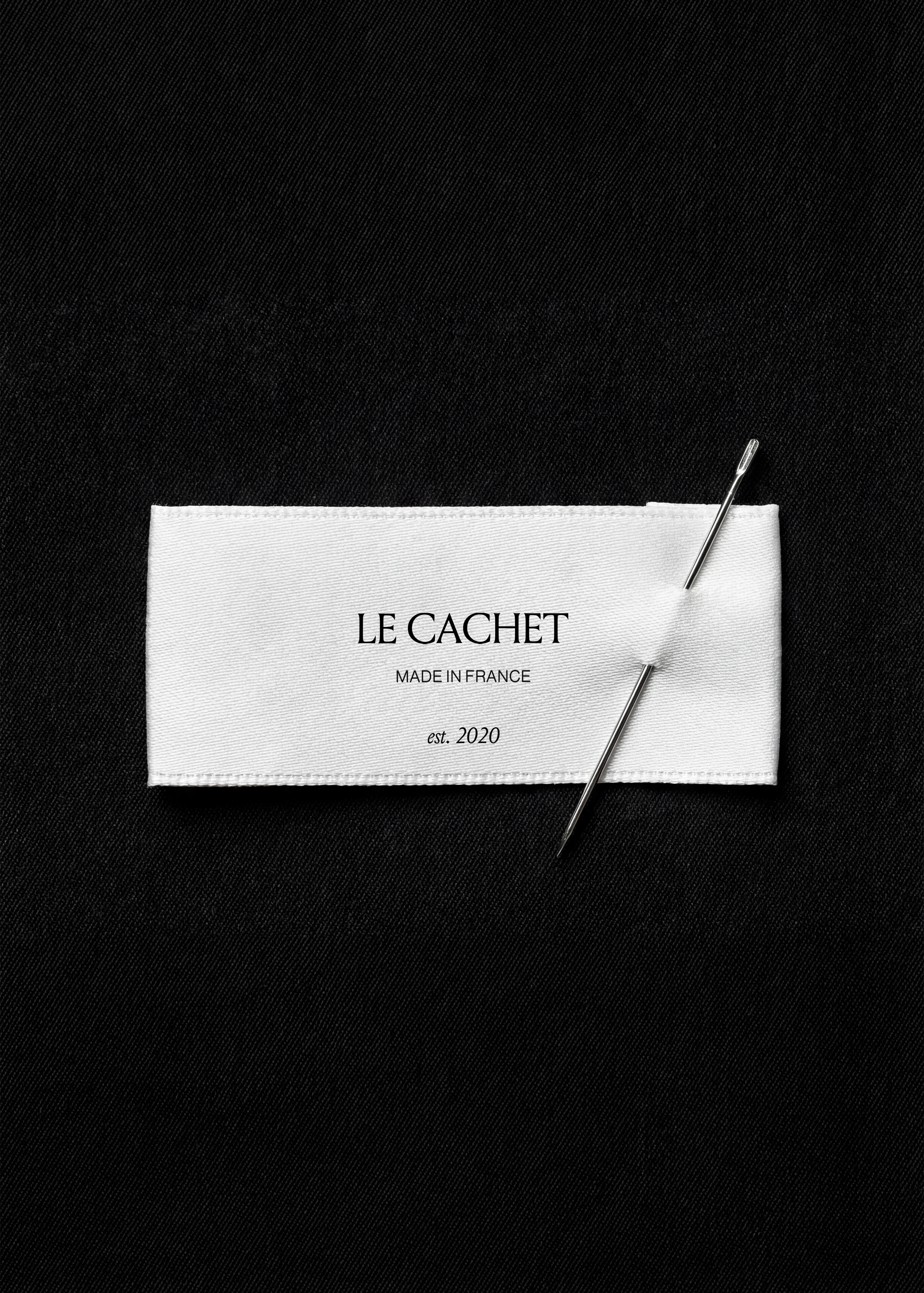 White satin garment label mockup on black fabric with a fine sewing needle threaded through, atelier still-life styling