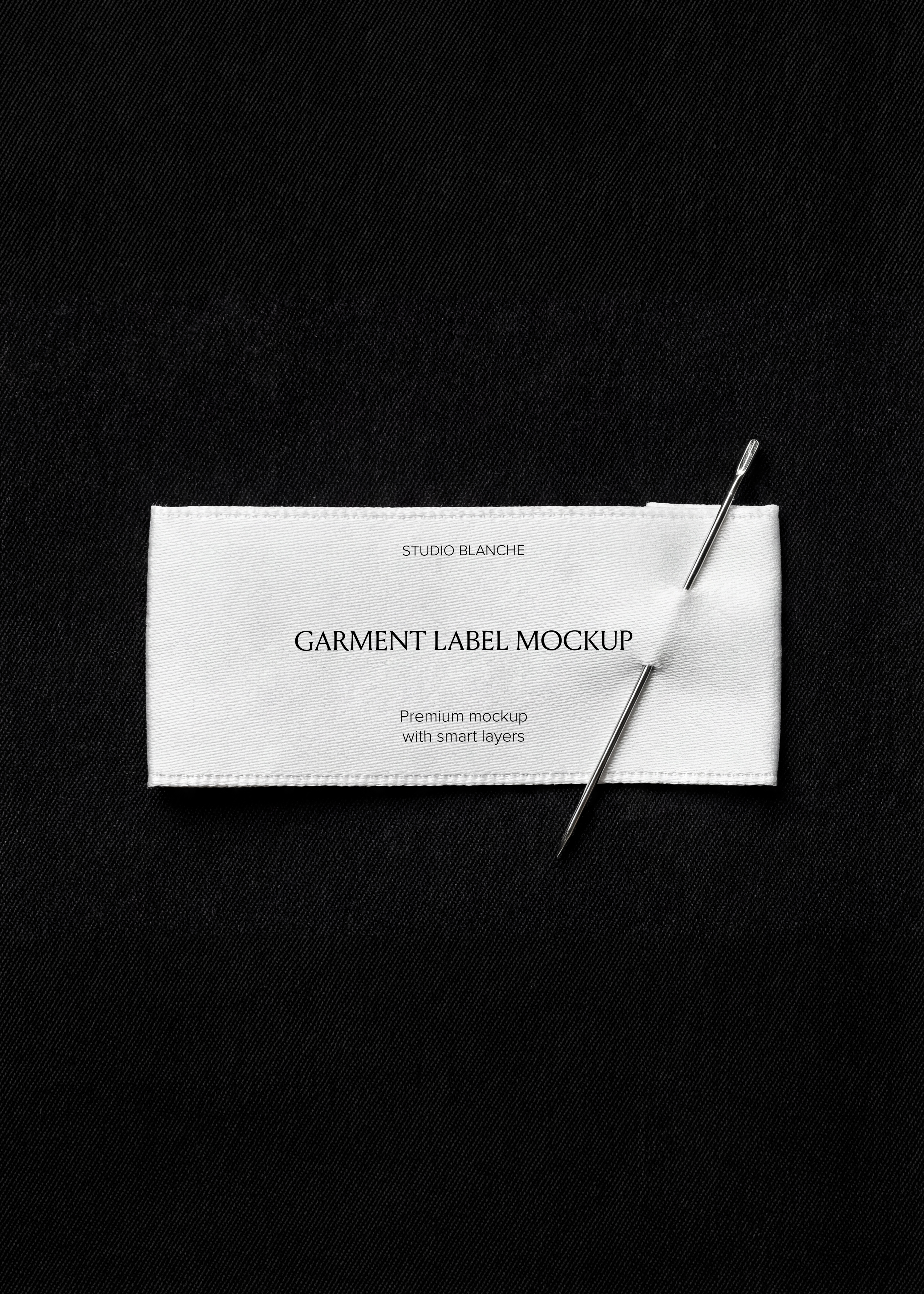 Woven garment label mockup pierced by a silver needle on dark twill, editorial fashion close-up in monochrome tones