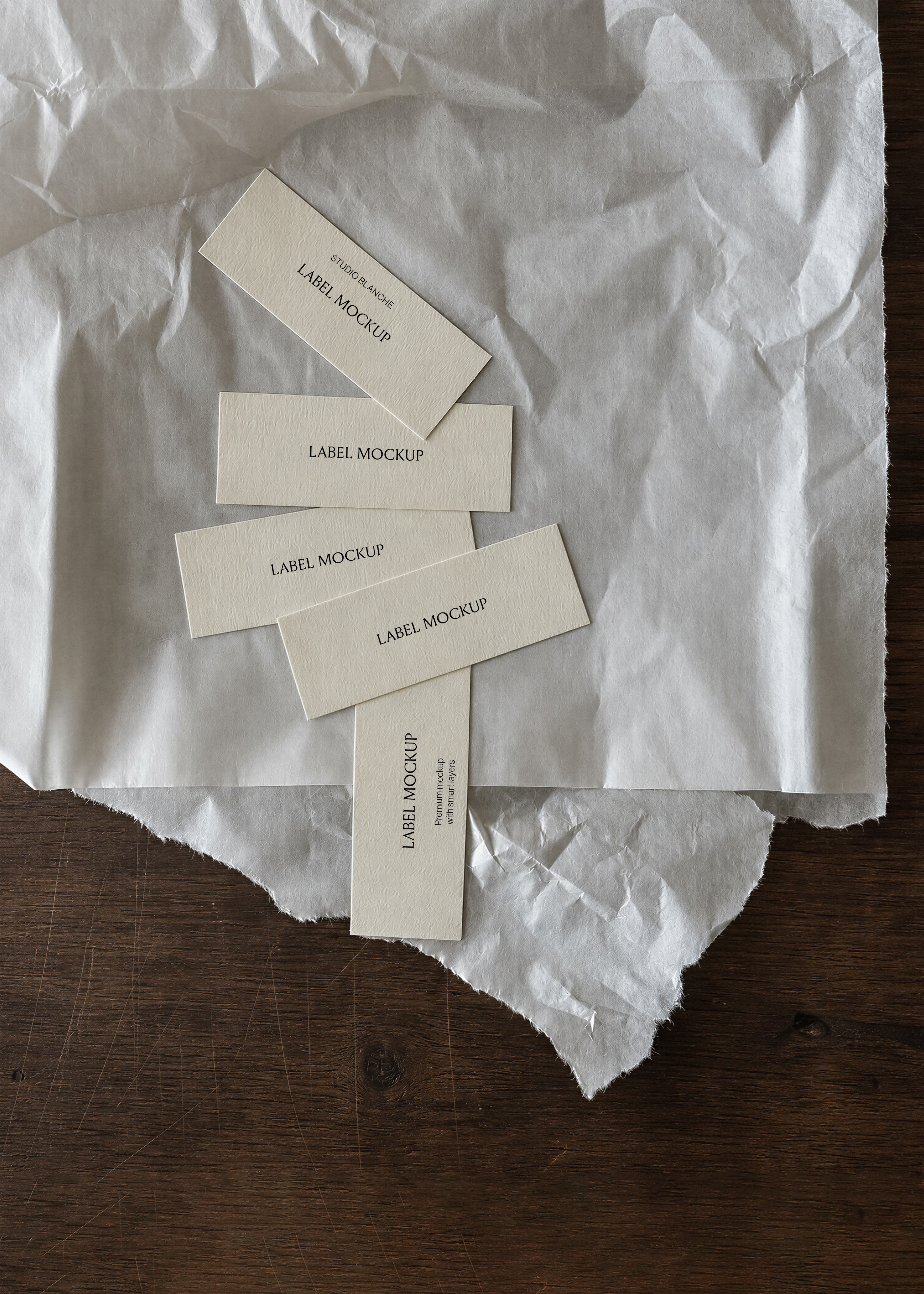 Paper label mockup, five cream labels cascading across white crumpled tissue paper on dark wood surface