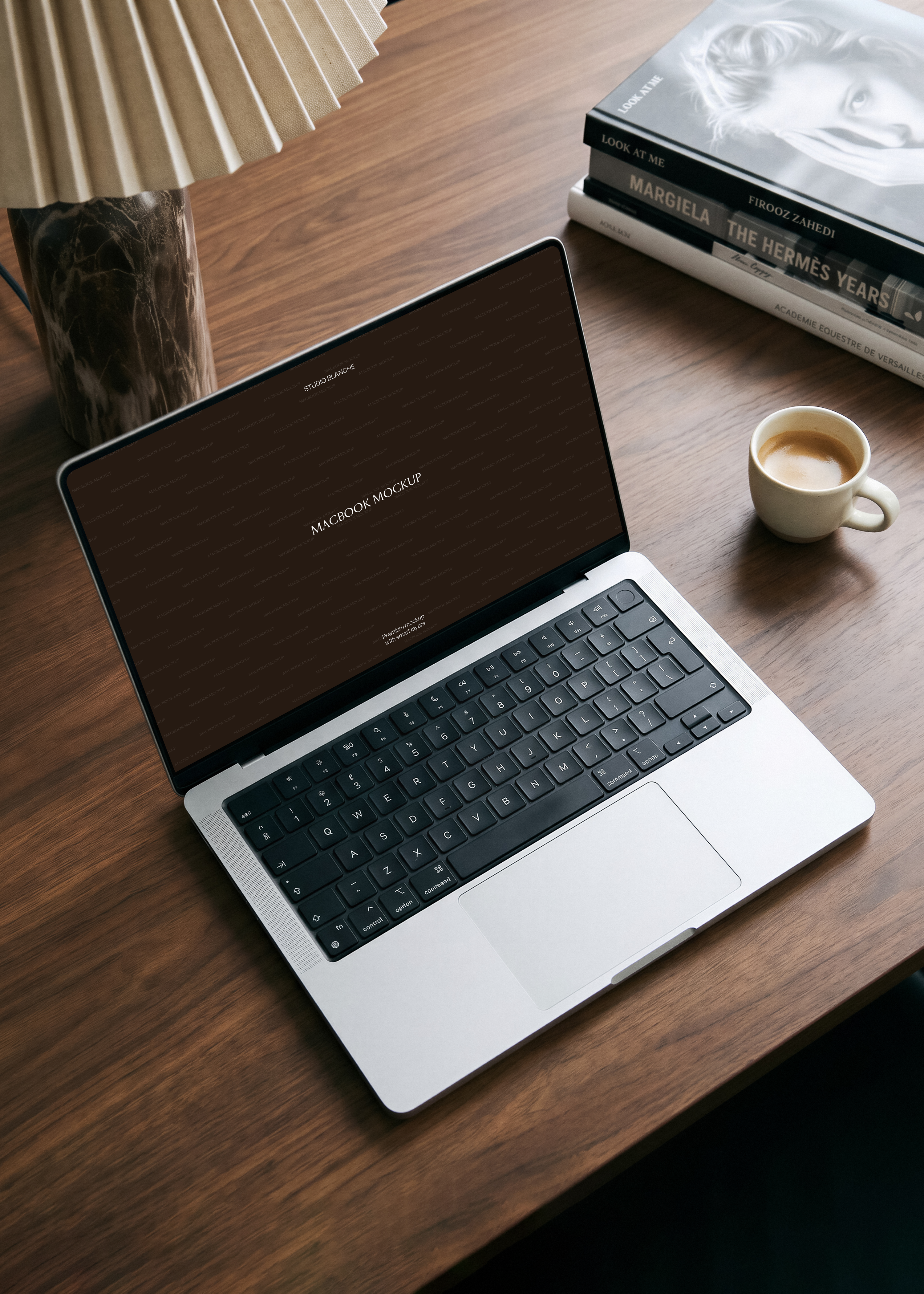 MacBook mockup from overhead on dark walnut desk with stacked fashion books, espresso cup, marble vase and pleated lamp shade