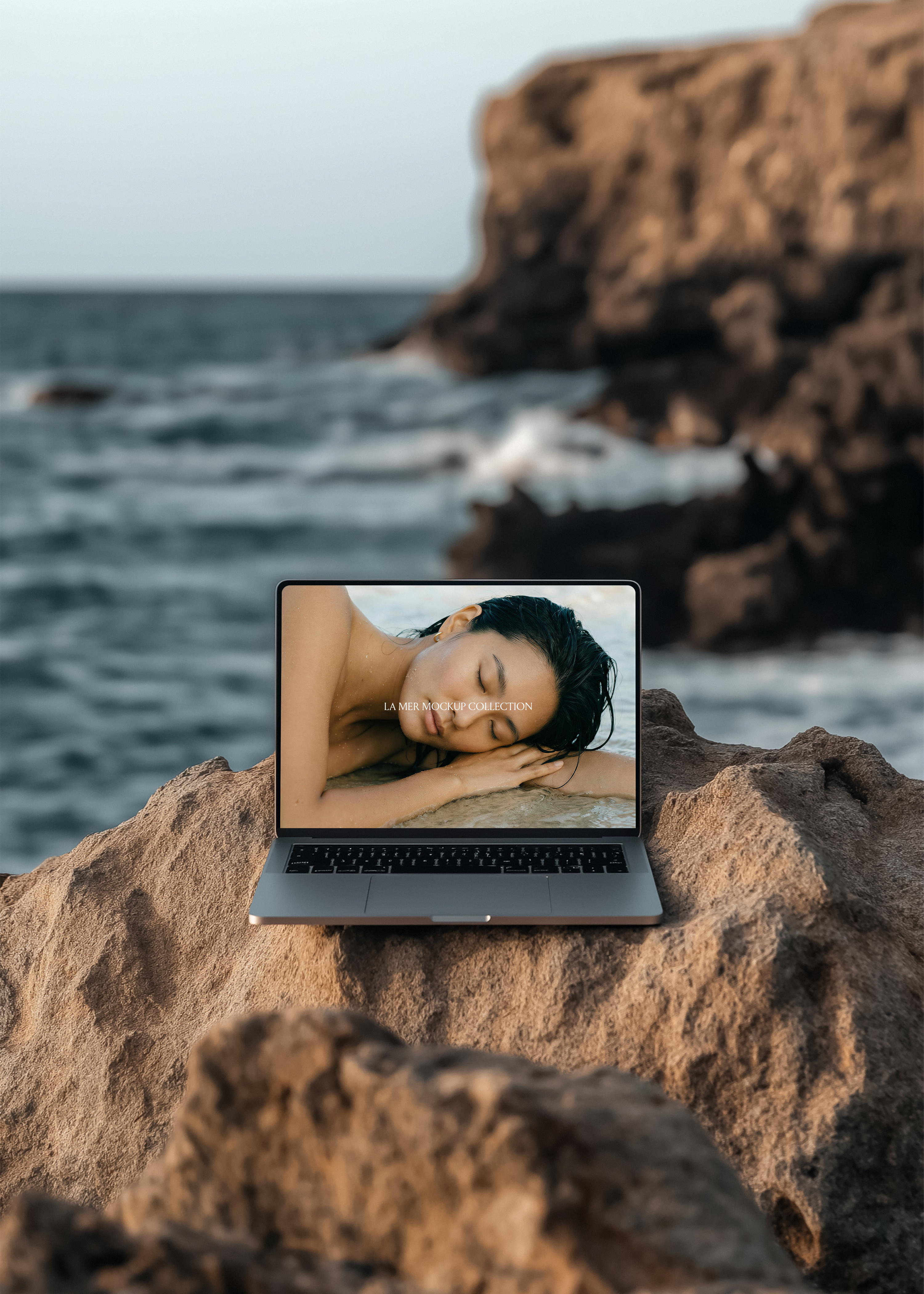 Silver MacBook open on volcanic rock, ocean and coastal cliffs blurred behind, warm dusk light, editorial scene