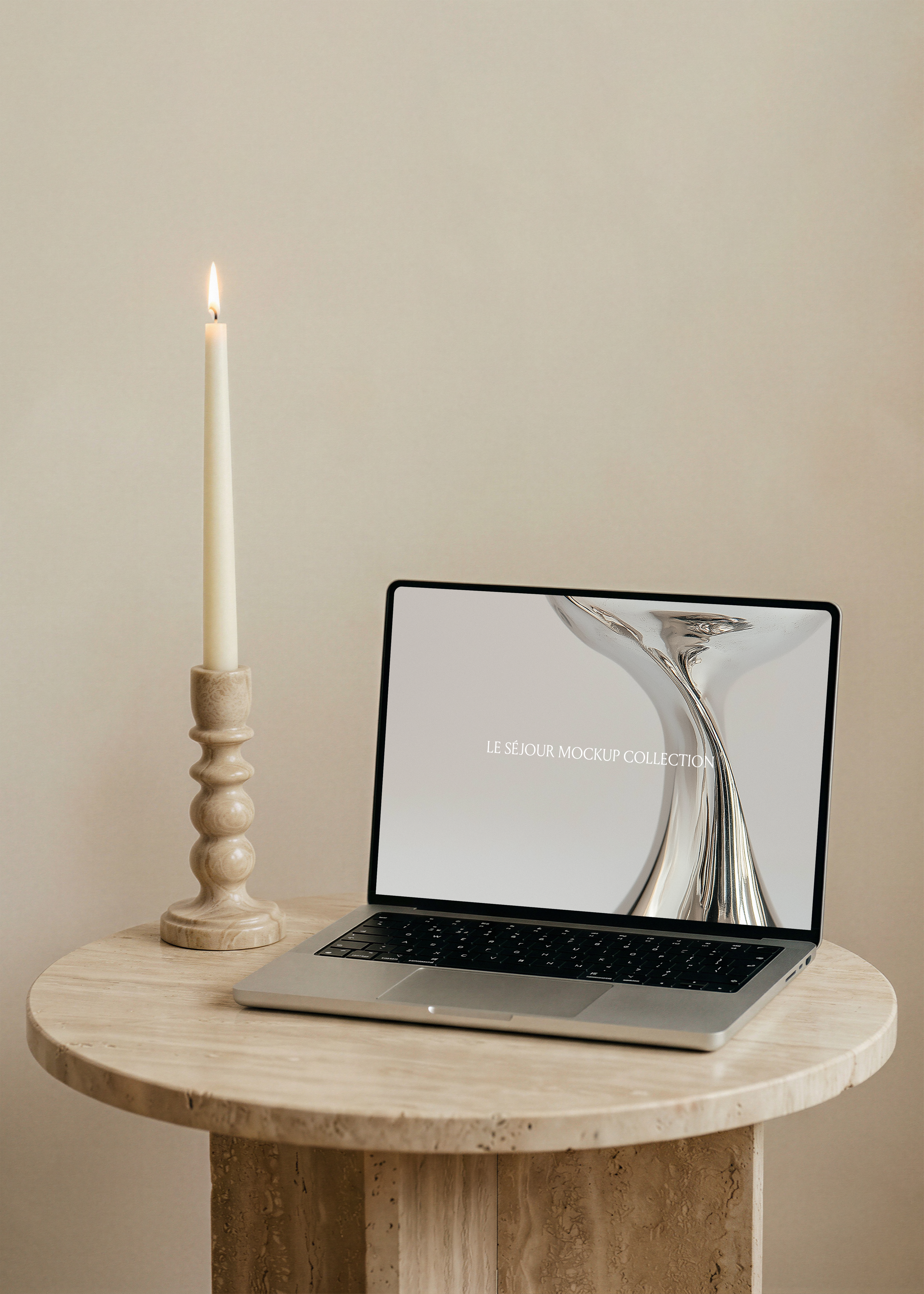 Open MacBook mockup on a round travertine side table beside a lit taper candle in a carved stone holder, warm beige wall