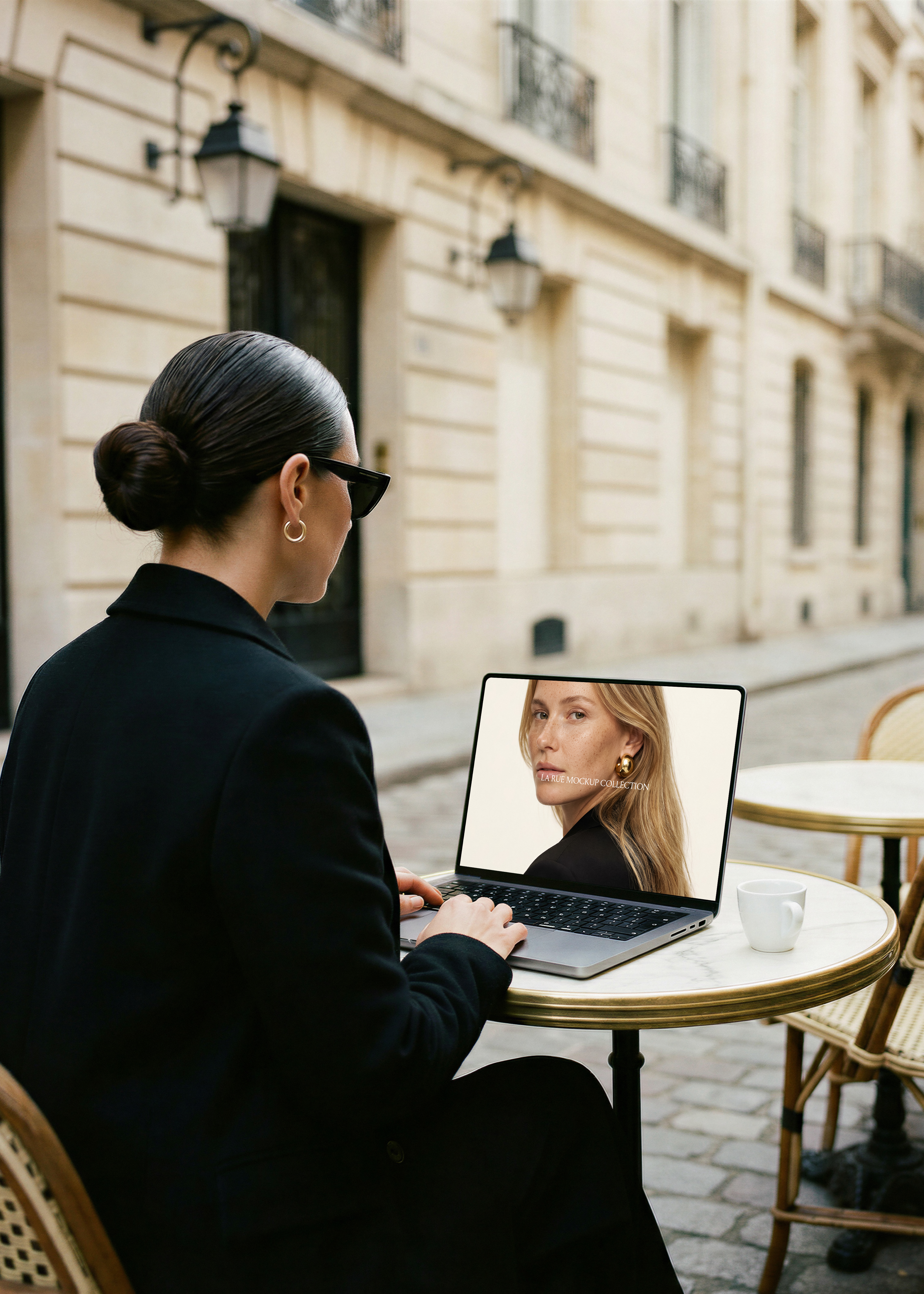 Woman in black at marble Paris café table with MacBook showing fashion website, Haussmann stone facade behind