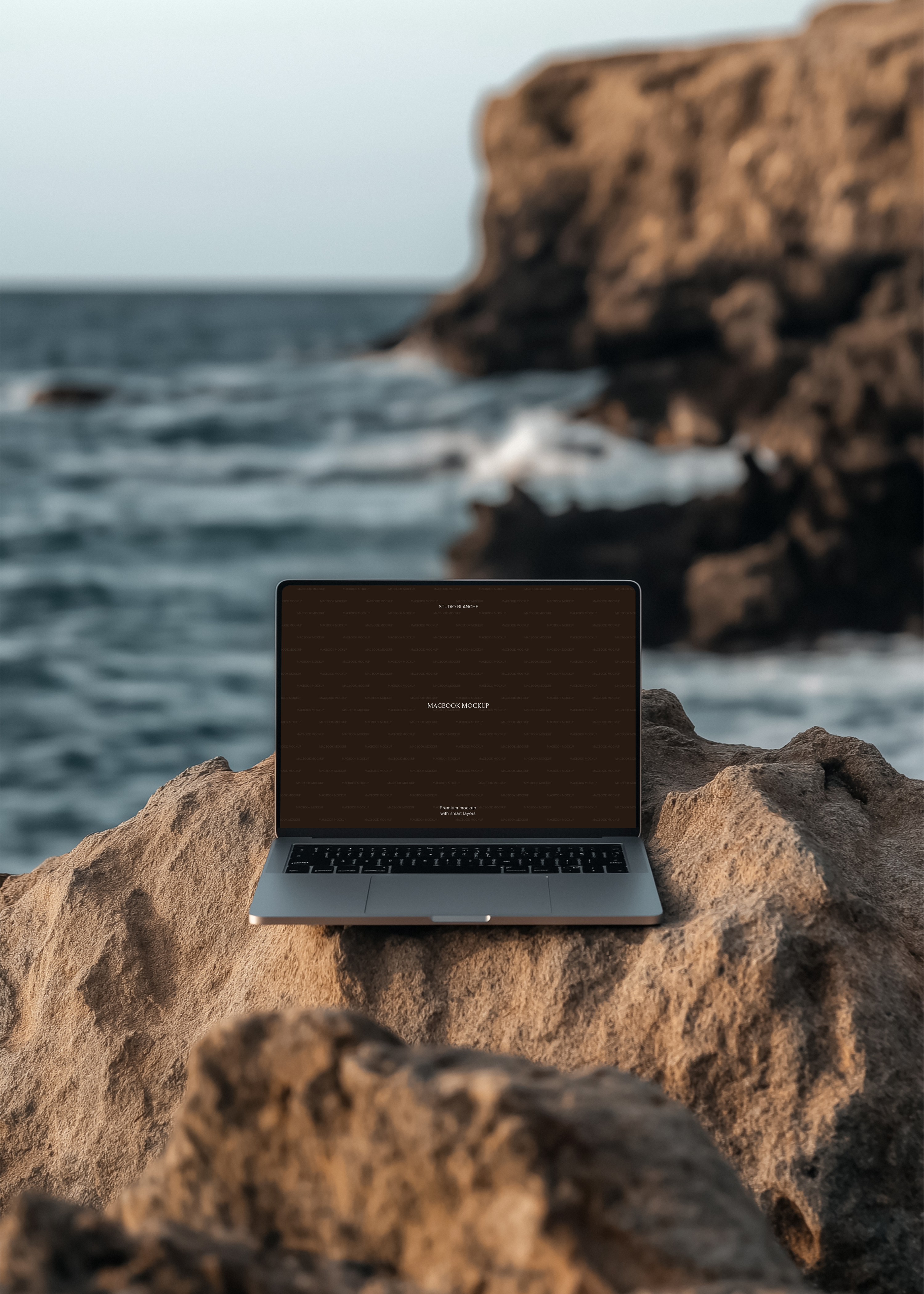 MacBook mockup on rocky coastal surface, dark placeholder screen, blurred sea cliffs and breaking waves in background