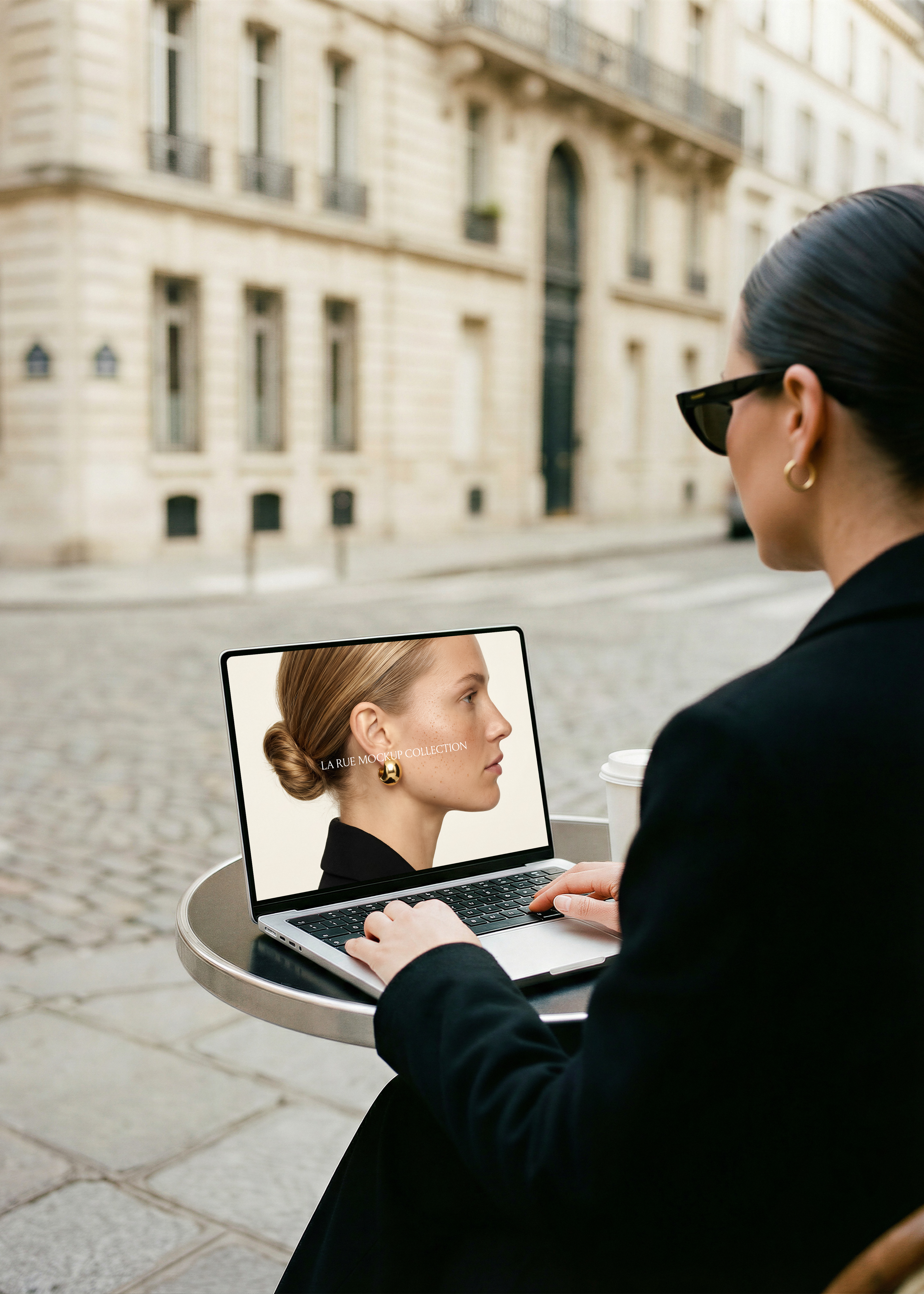 Woman in black seated at Paris street terrace with MacBook showing editorial fashion website, cobblestone background