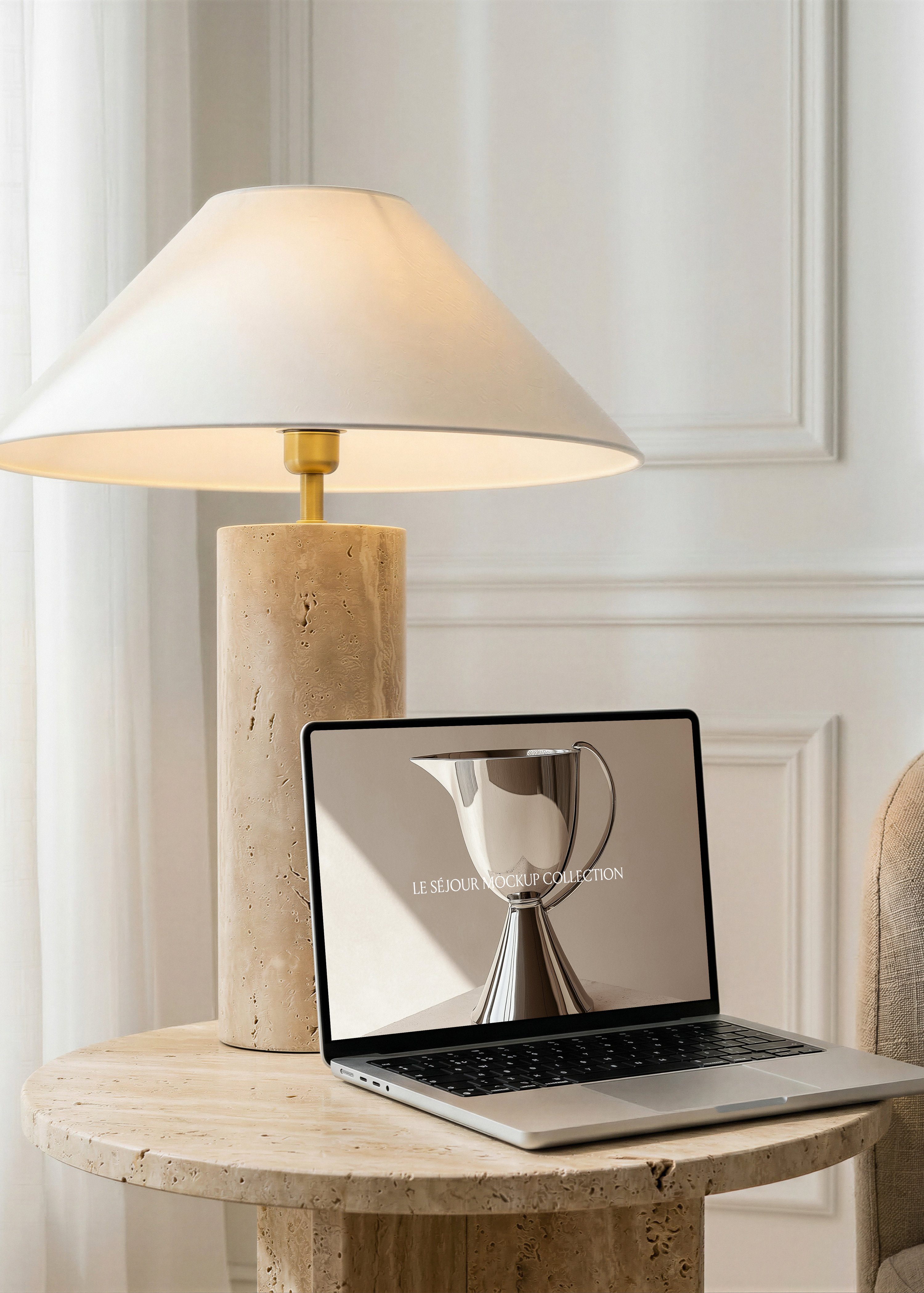 Open MacBook mockup on a round travertine side table beside a travertine lamp with linen shade, white panelled wall behind