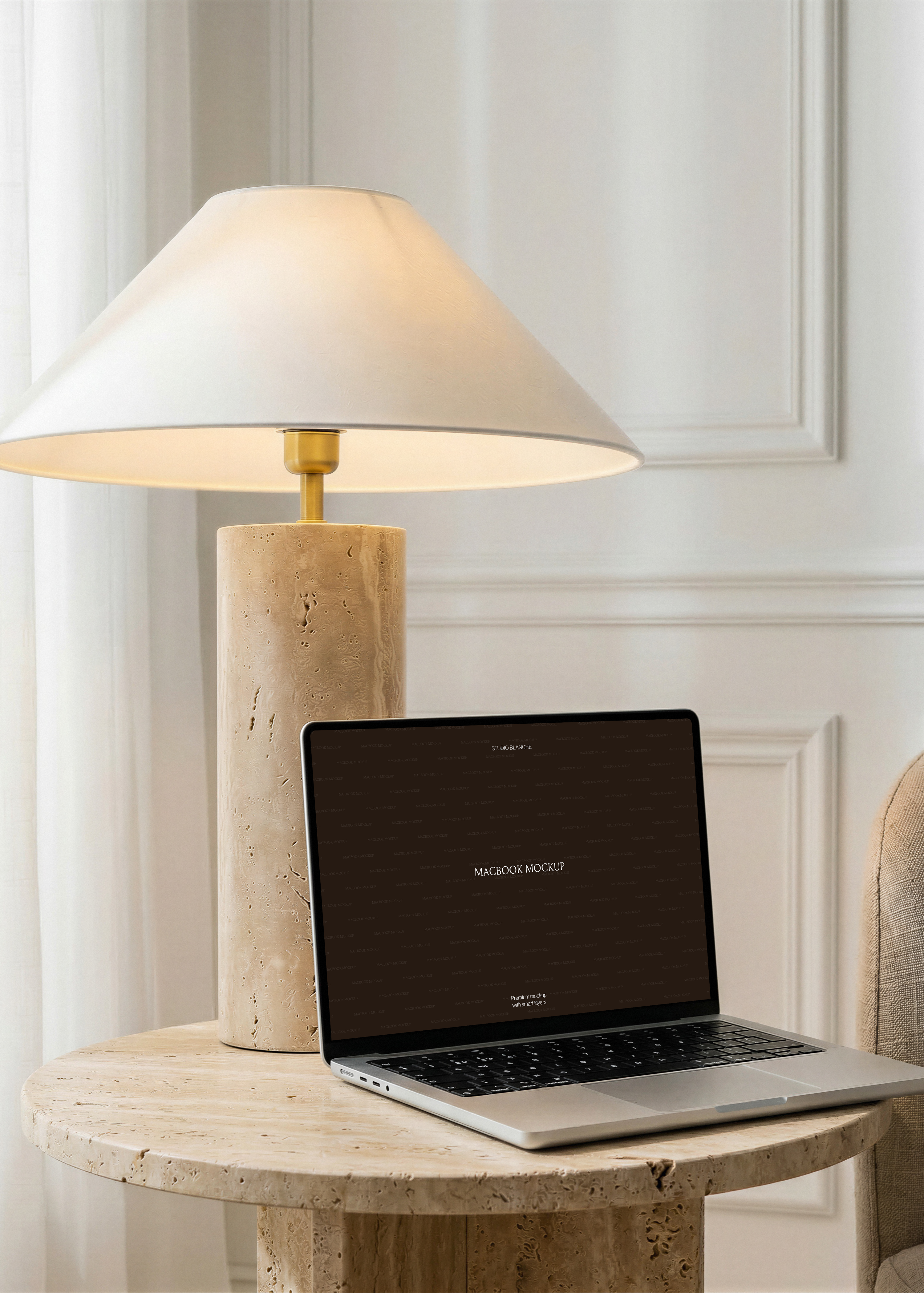 MacBook mockup detail of a dark laptop screen angled beside a pitted travertine lamp base, linen armchair edge in warm light
