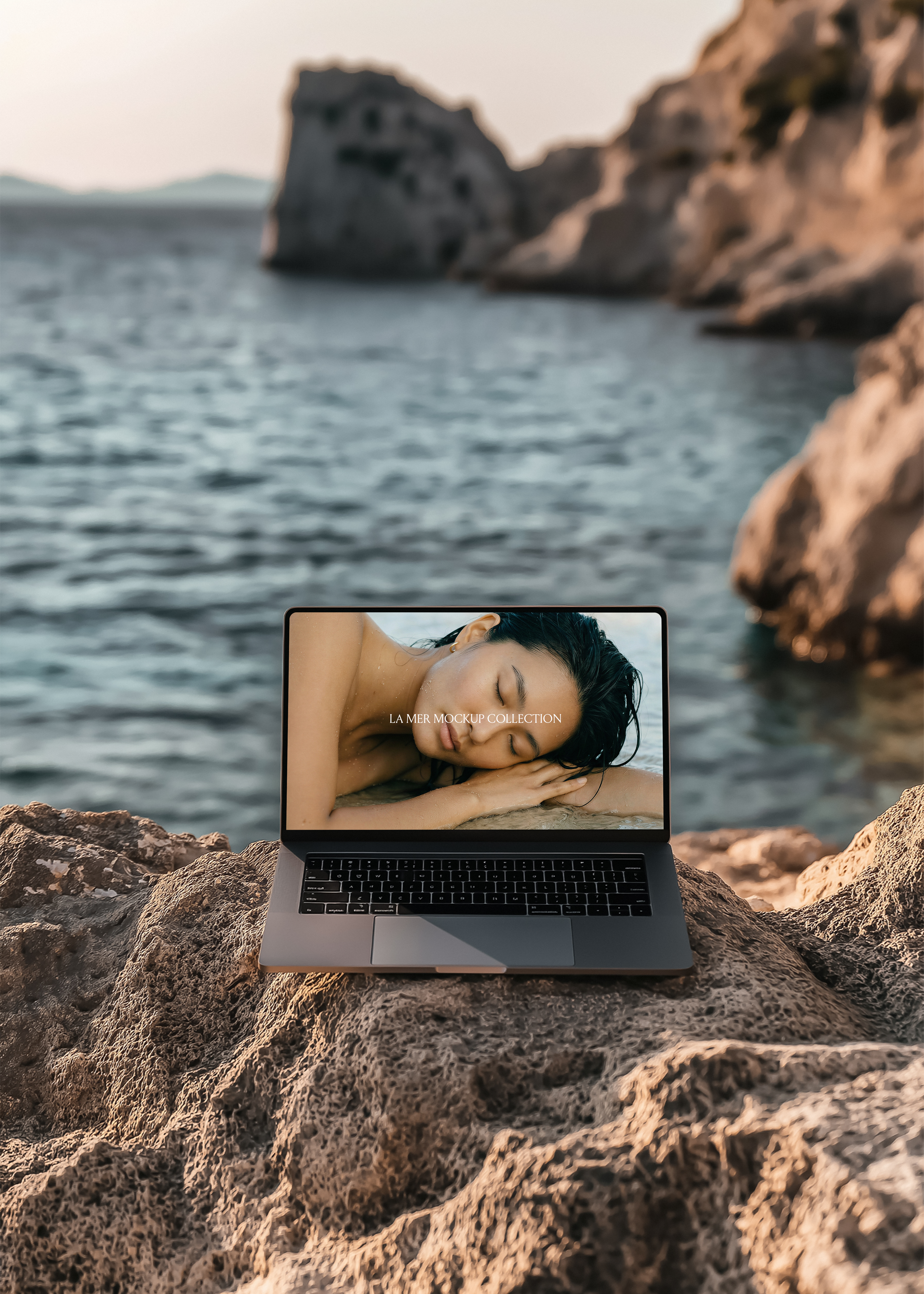 Silver MacBook open on warm sun-lit limestone rock, wide coastal bay and dramatic sea arch cliff blurred softly behind