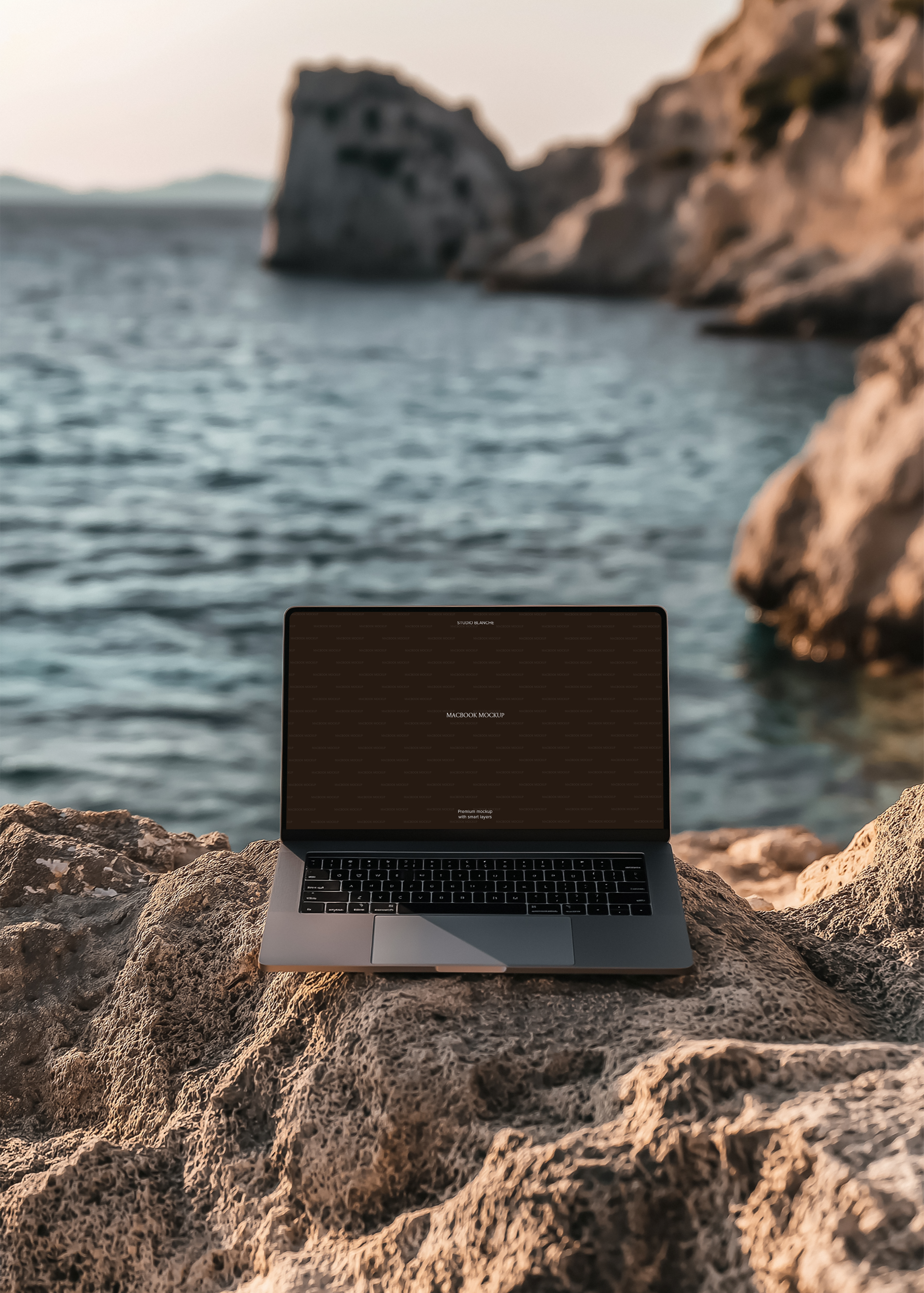 MacBook mockup with dark placeholder screen on bleached coastal rock, calm blue bay and rocky headland in background