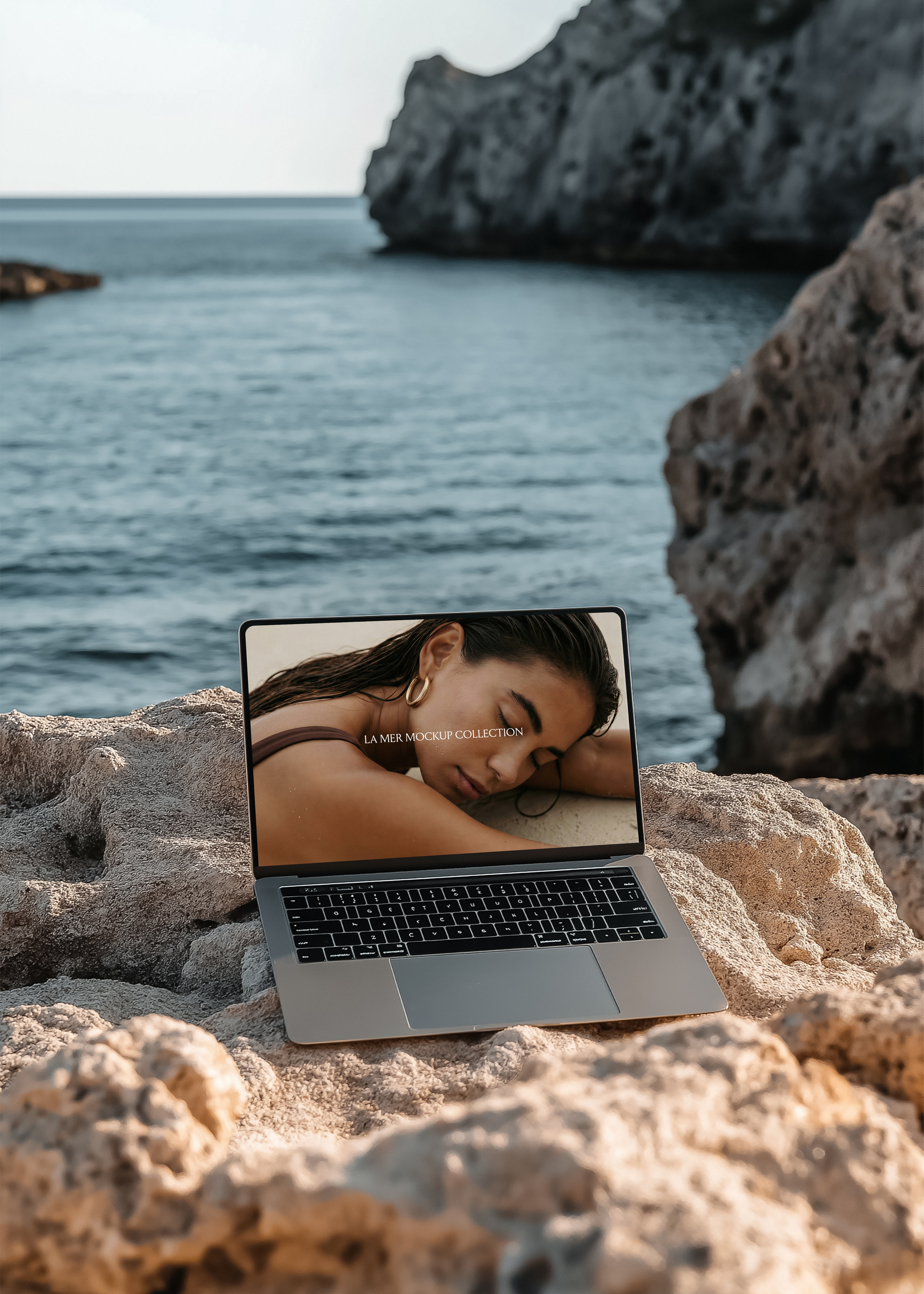 Silver MacBook open on textured coastal rock, warm golden hour light, calm cove and limestone sea stacks blurred behind